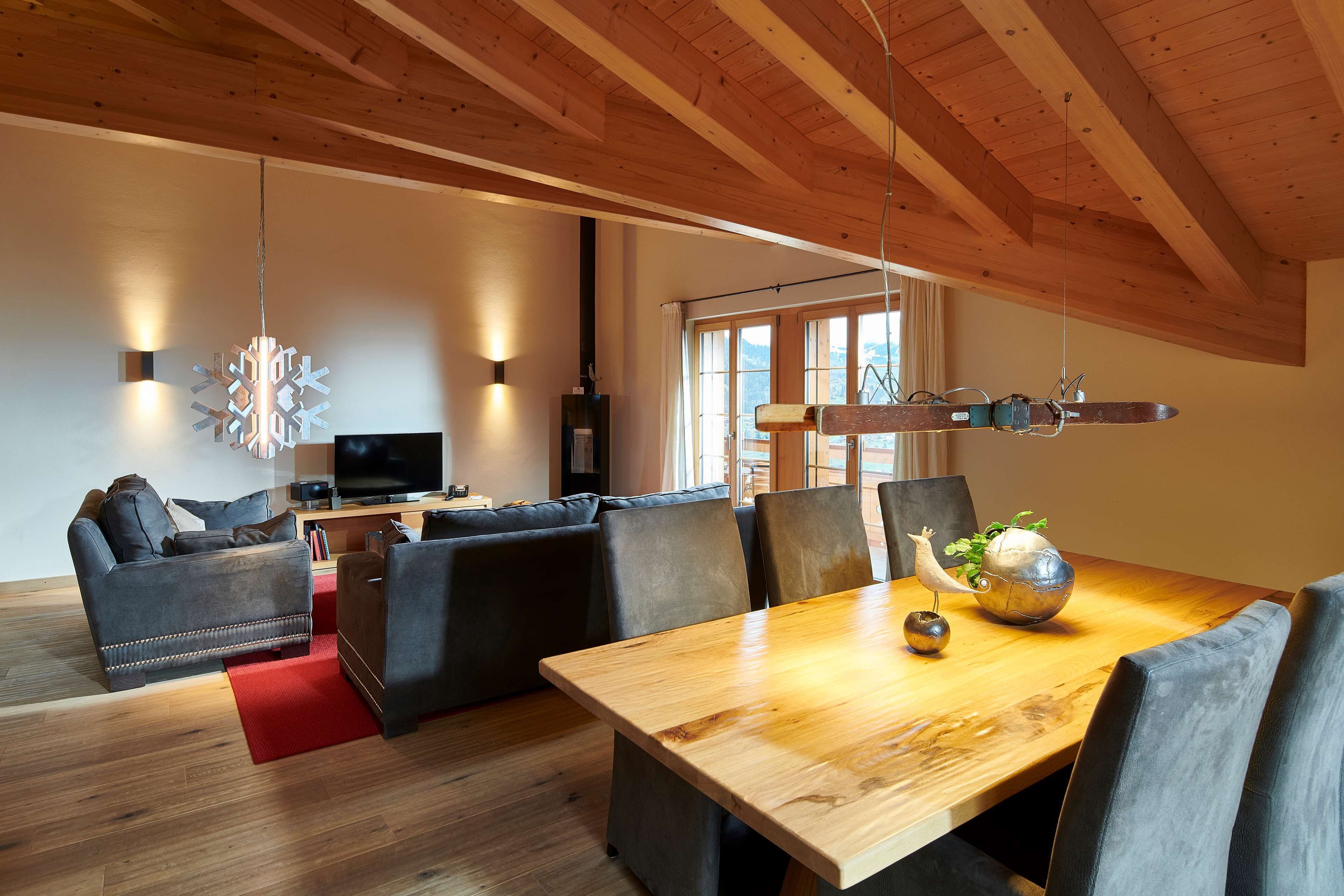 A modern living room with a wooden ceiling, a dining table with chairs, a sofa set, and a television.