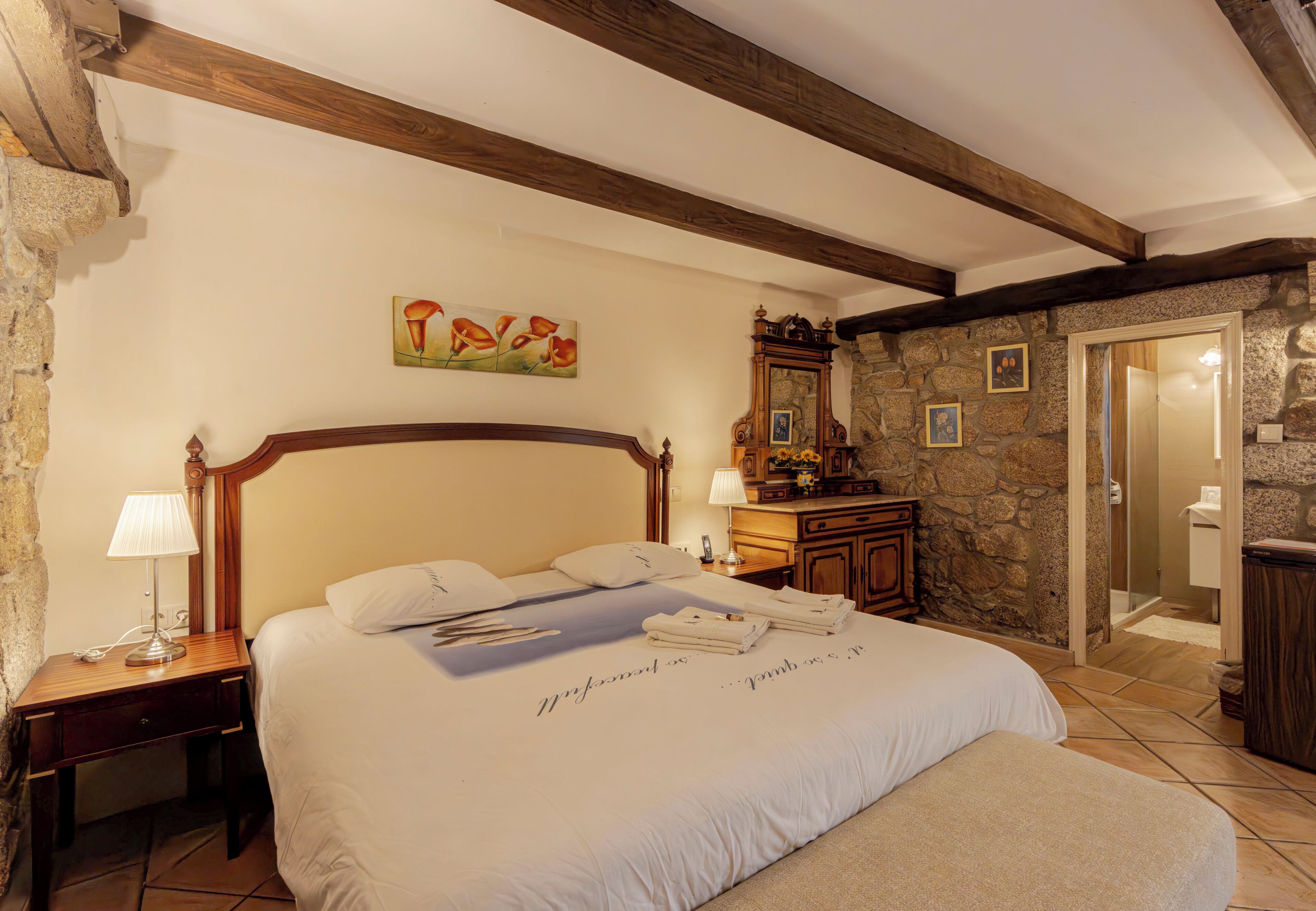 A bedroom with a large bed, wooden headboard, stone wall, and a small desk with a lamp.