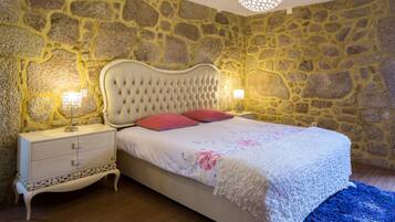 Quarto com parede de pedra, cabeceira estofada, mesa de cabeceira branca e tapete azul.