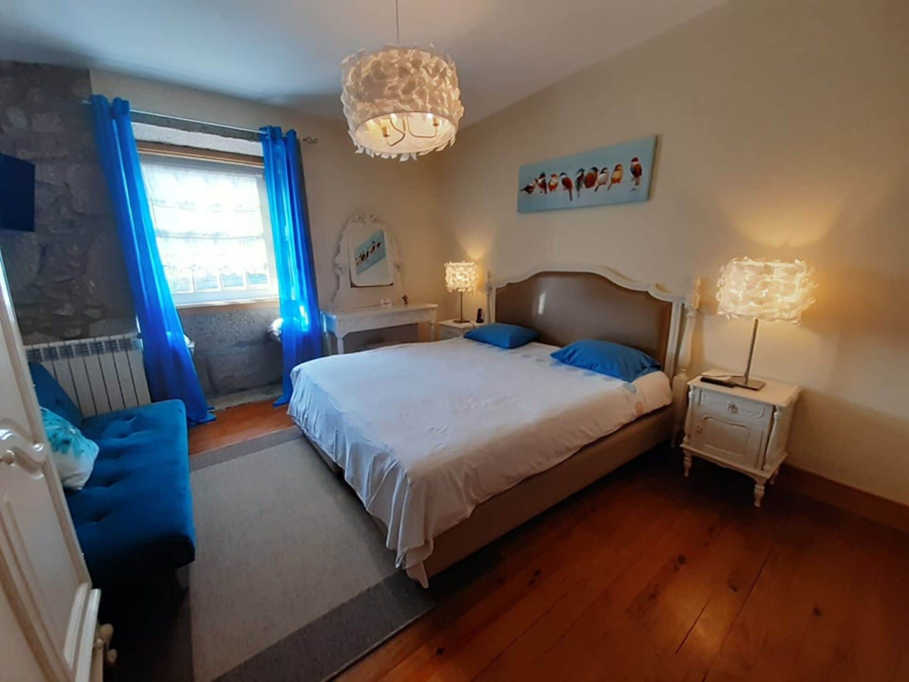 Ein Schlafzimmer mit einem großen Bett, zwei Nachttischen, einer Kommode, einem Fenster mit blauen Vorhängen und einer Wandleuchte.