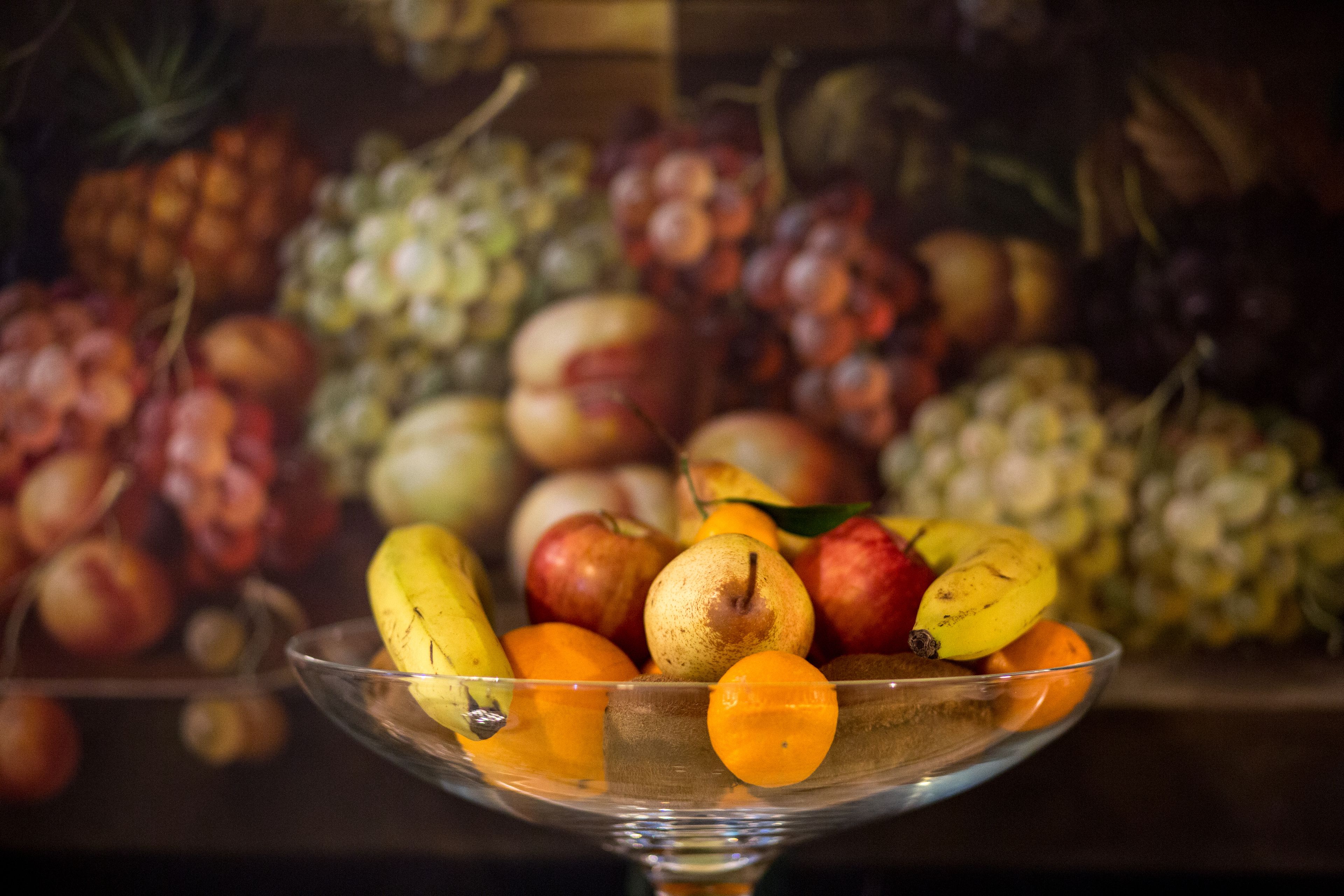 Een glazen schaal gevuld met diverse soorten fruit, zoals bananen, appels en sinaasappels, voor een onscherpe achtergrond van nog meer fruit.