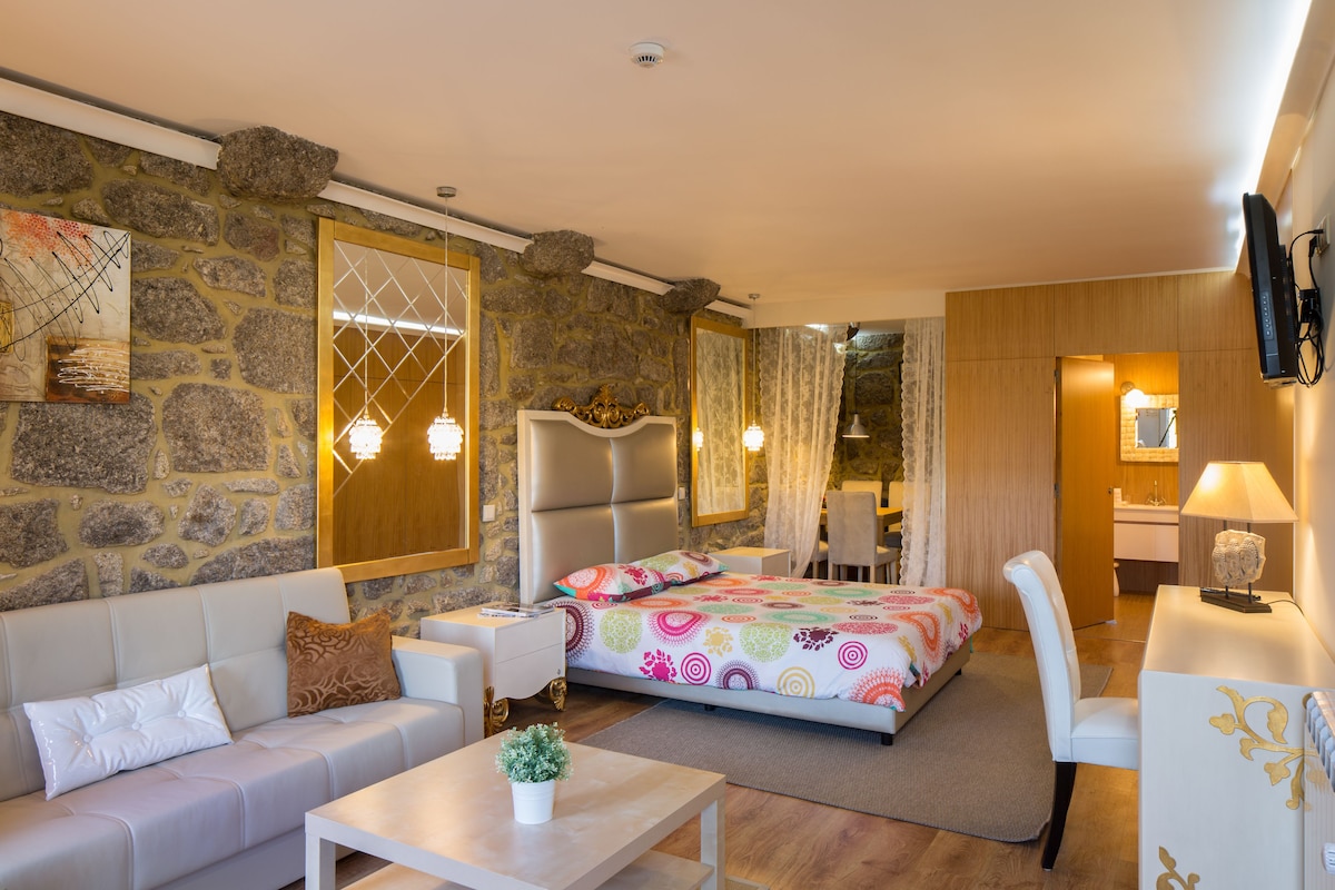 A modern hotel room with a stone accent wall, a large bed with colorful bedding, a sofa, a desk, and a chair.