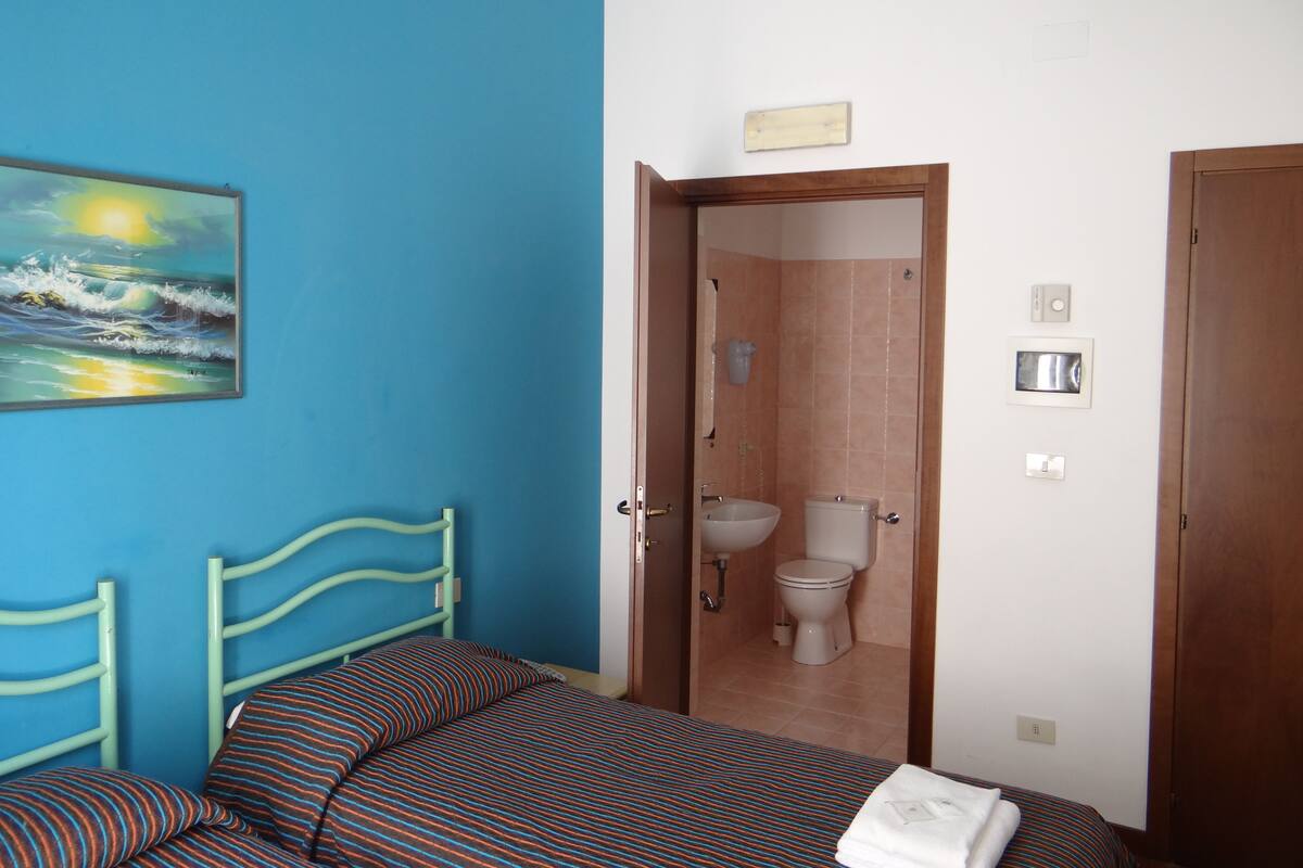 A bedroom with a bed, a painting of a sunset, a bathroom with a toilet and sink, and a wooden door.