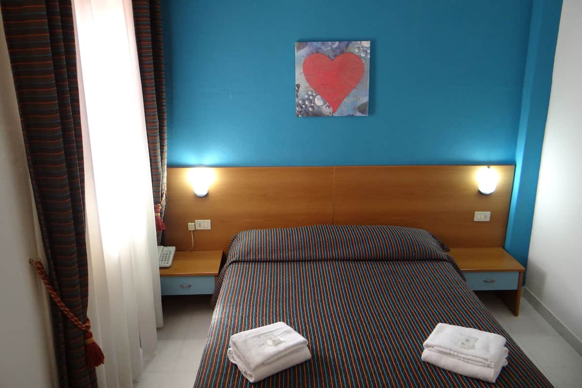 A hotel room with a bed, a headboard with lights, a wall with a heart-shaped painting, and a bedside table with a phone.
