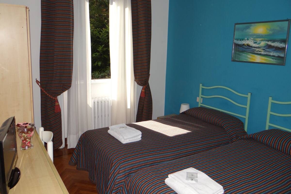 A bedroom with two beds, a window with curtains, a painting on the wall, and a bedside table with a lamp.