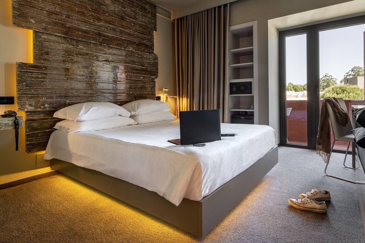 A modern hotel room with a large bed, a laptop on the bed, a desk with a chair, and a view of the outdoors through a sliding door.