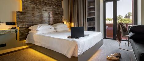 A modern hotel room with a large bed, a laptop on the bed, a desk with a chair, and a view of the outdoors through a sliding door.