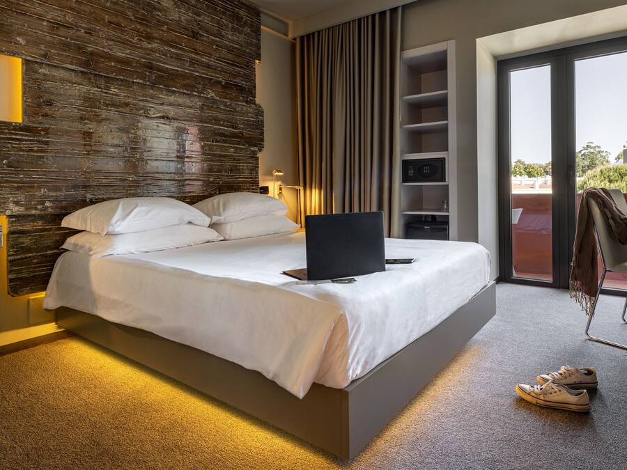 A modern hotel room with a large bed, a laptop on the bed, a desk with a chair, and a view of the outdoors through a sliding door.