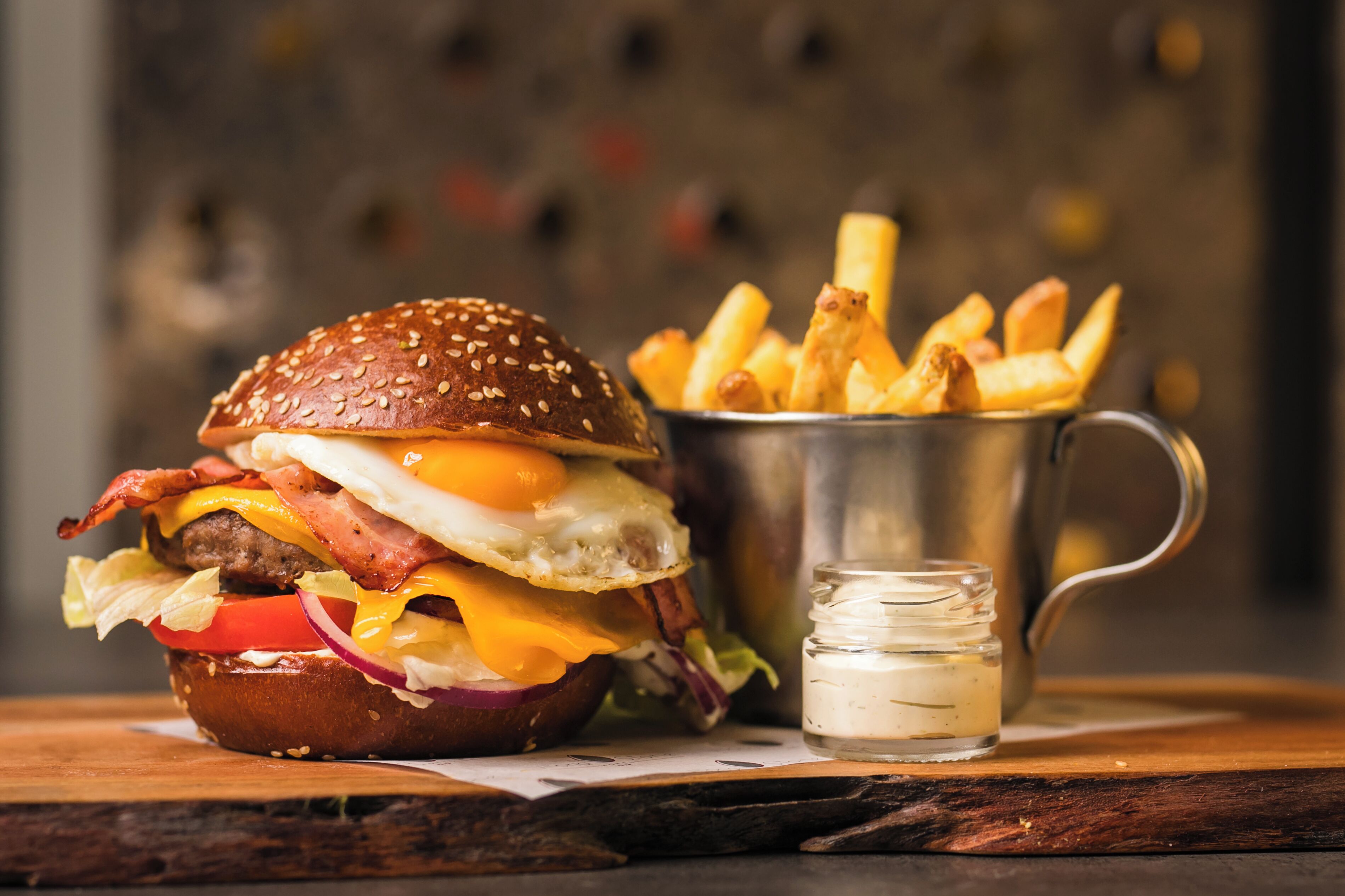 A burger with fries and a small jar of sauce on a wooden table.