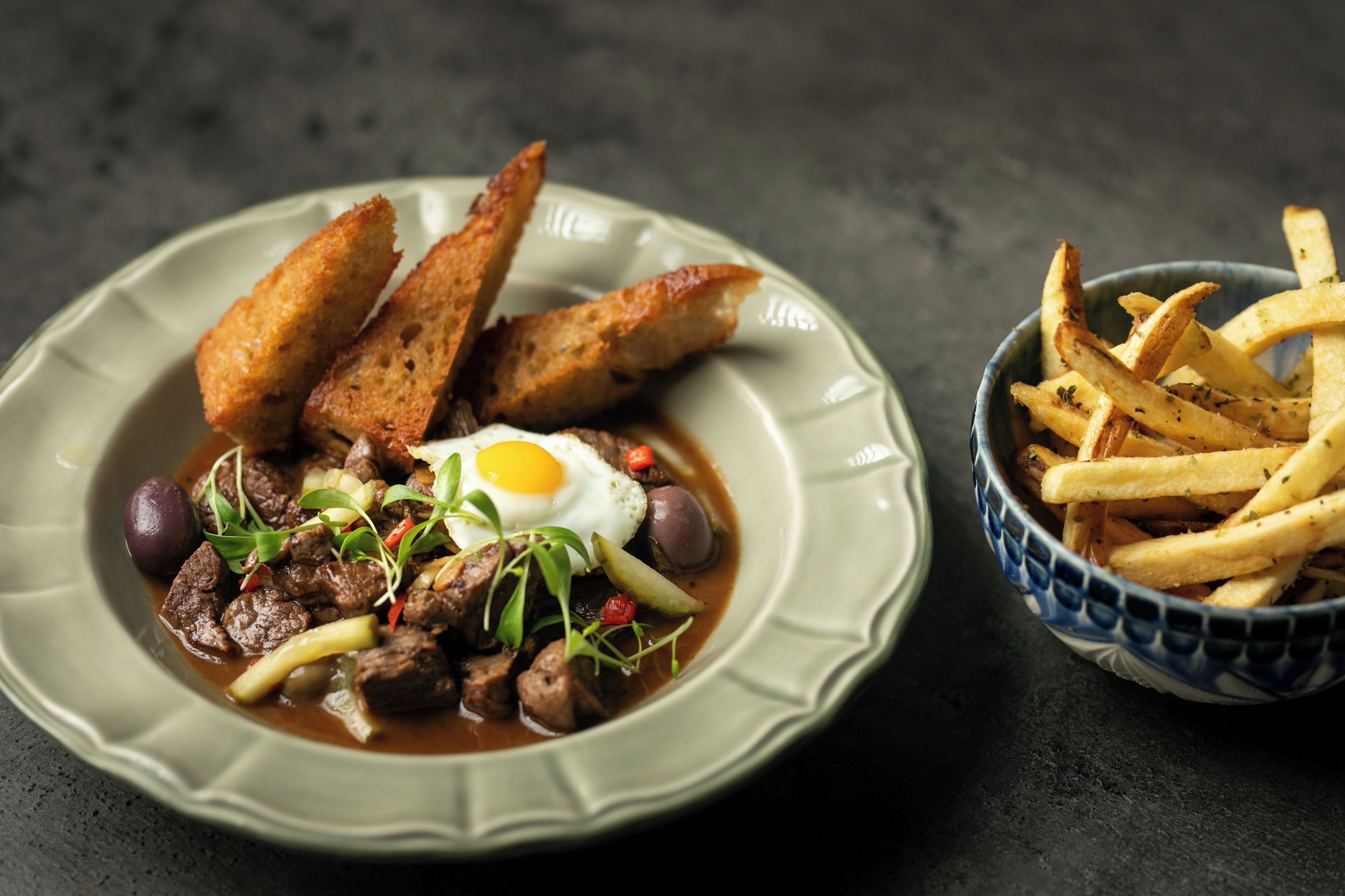 A plate of beef stew with egg and vegetables, accompanied by toasted bread and French fries.