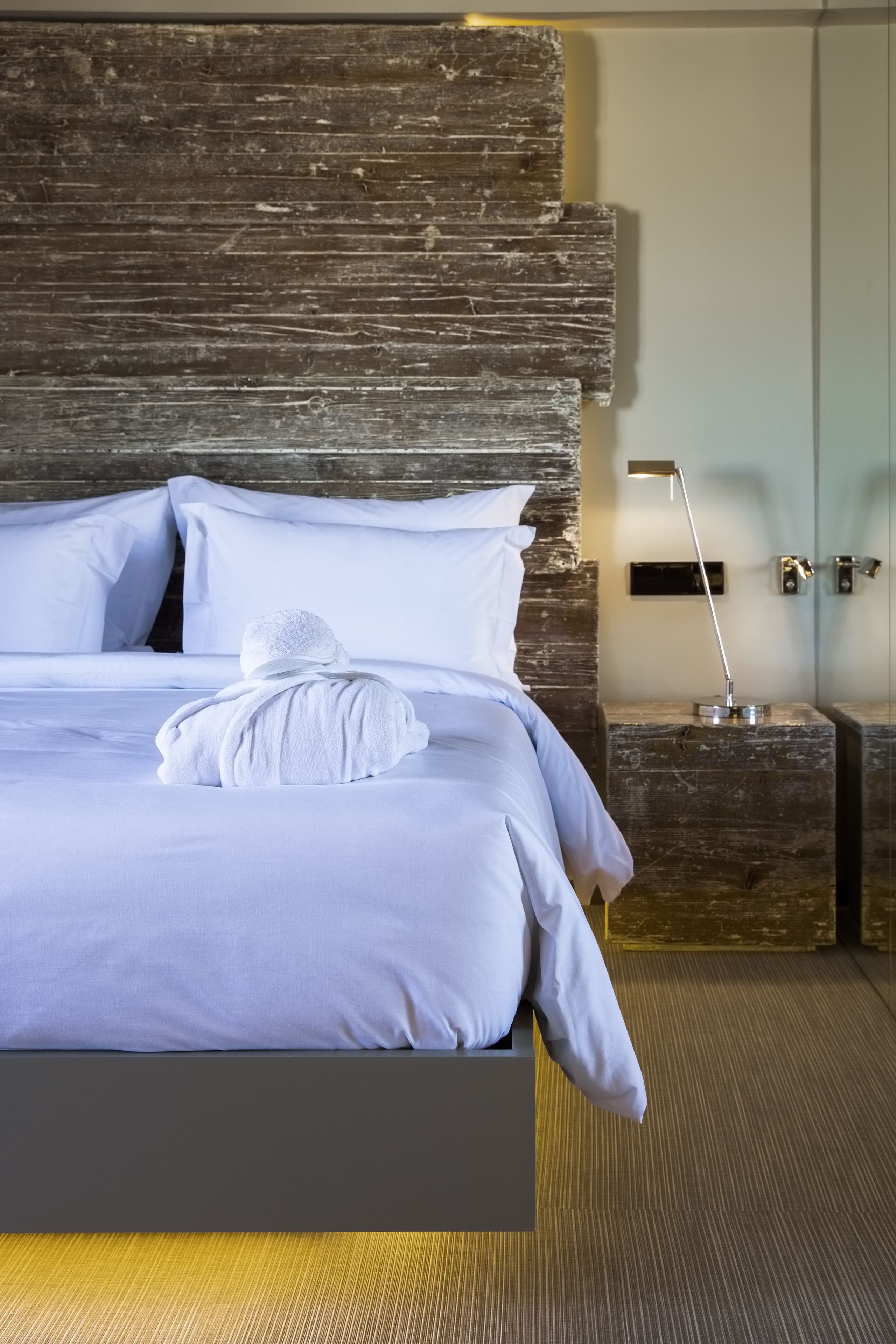Una cama bien hecha con sábanas y toalla blancos, un cabecero moderno, una mesita de noche con una lámpara y una pared decorativa de madera.
