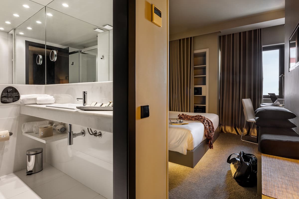 A modern hotel room with a bathroom, a bed, a desk, and a window.