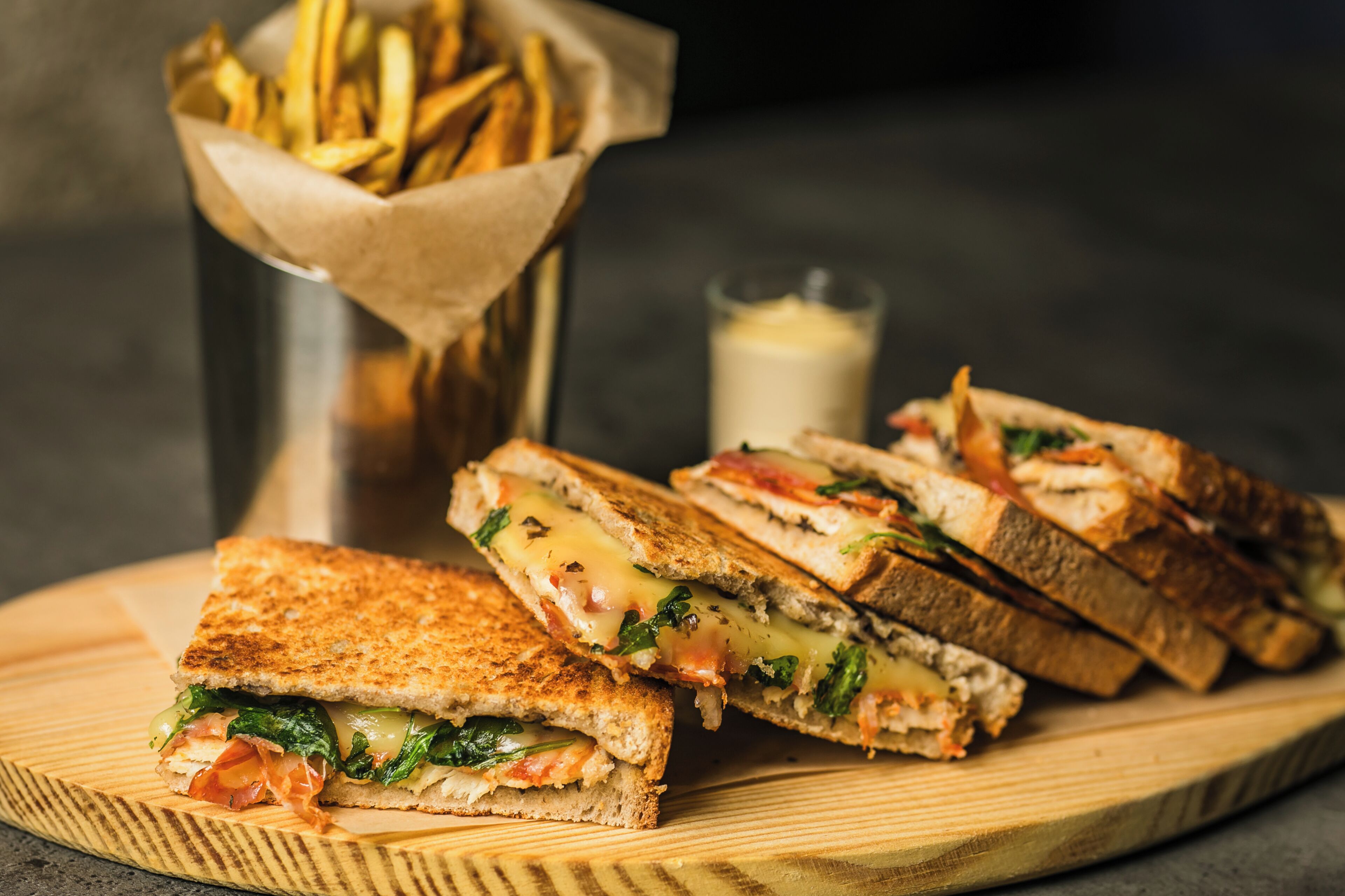 Un sándwich cortado por la mitad sobre una tabla de madera, con papas fritas en una cesta y un vaso de leche al fondo.