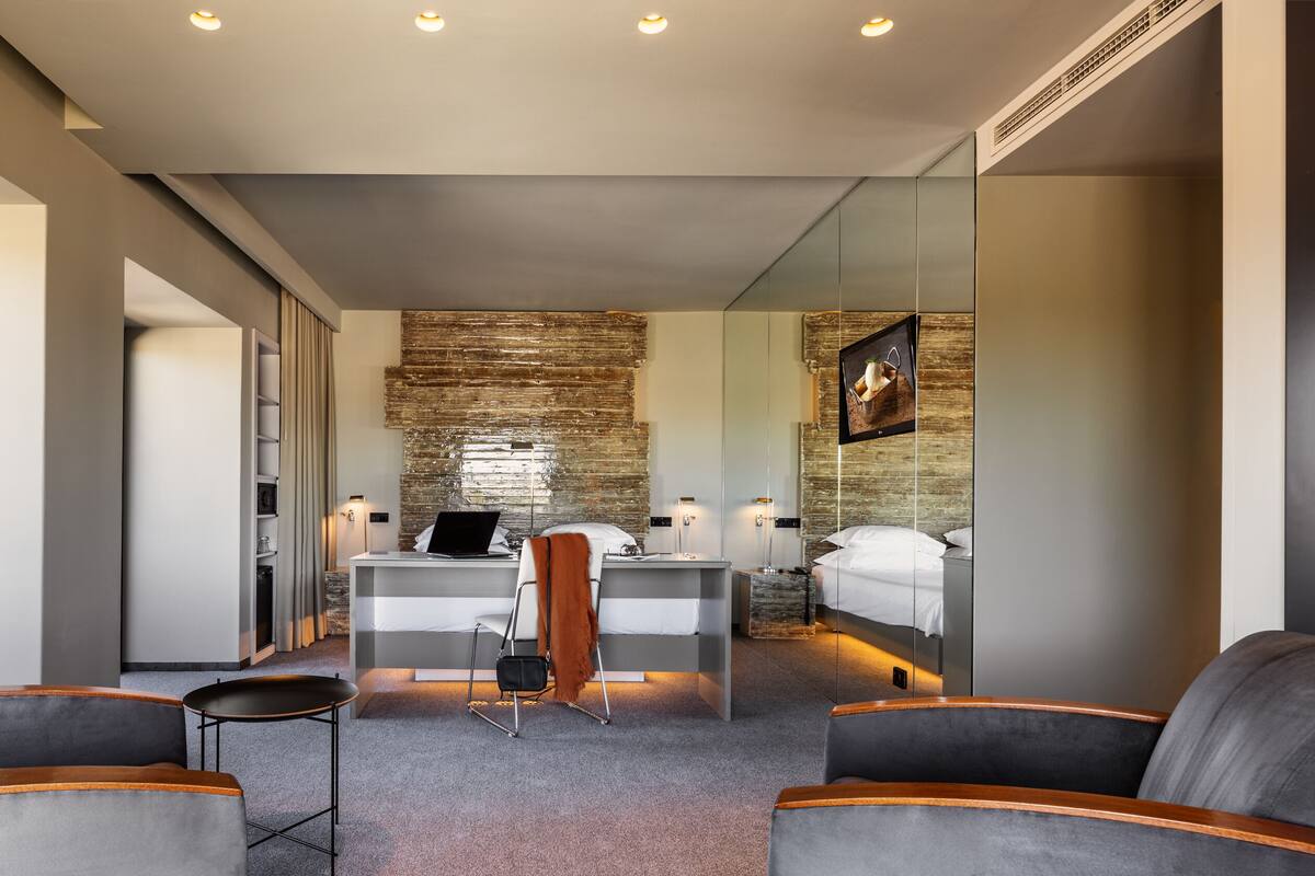 A modern hotel room with a large mirror, a desk with a laptop, a bed, and two armchairs.