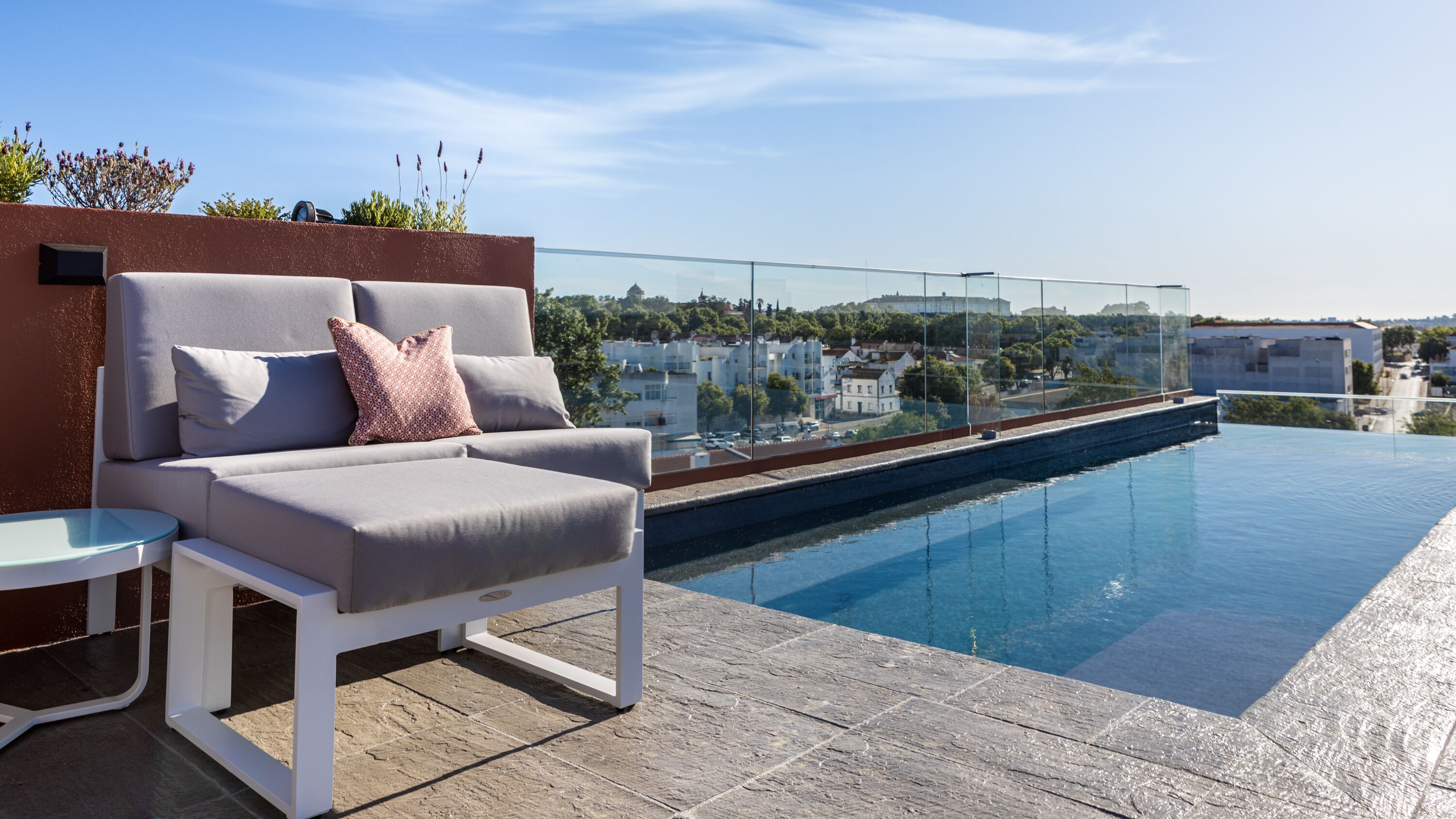 A modern outdoor seating area with a pool, clear glass balustrade, and a view of a residential area.