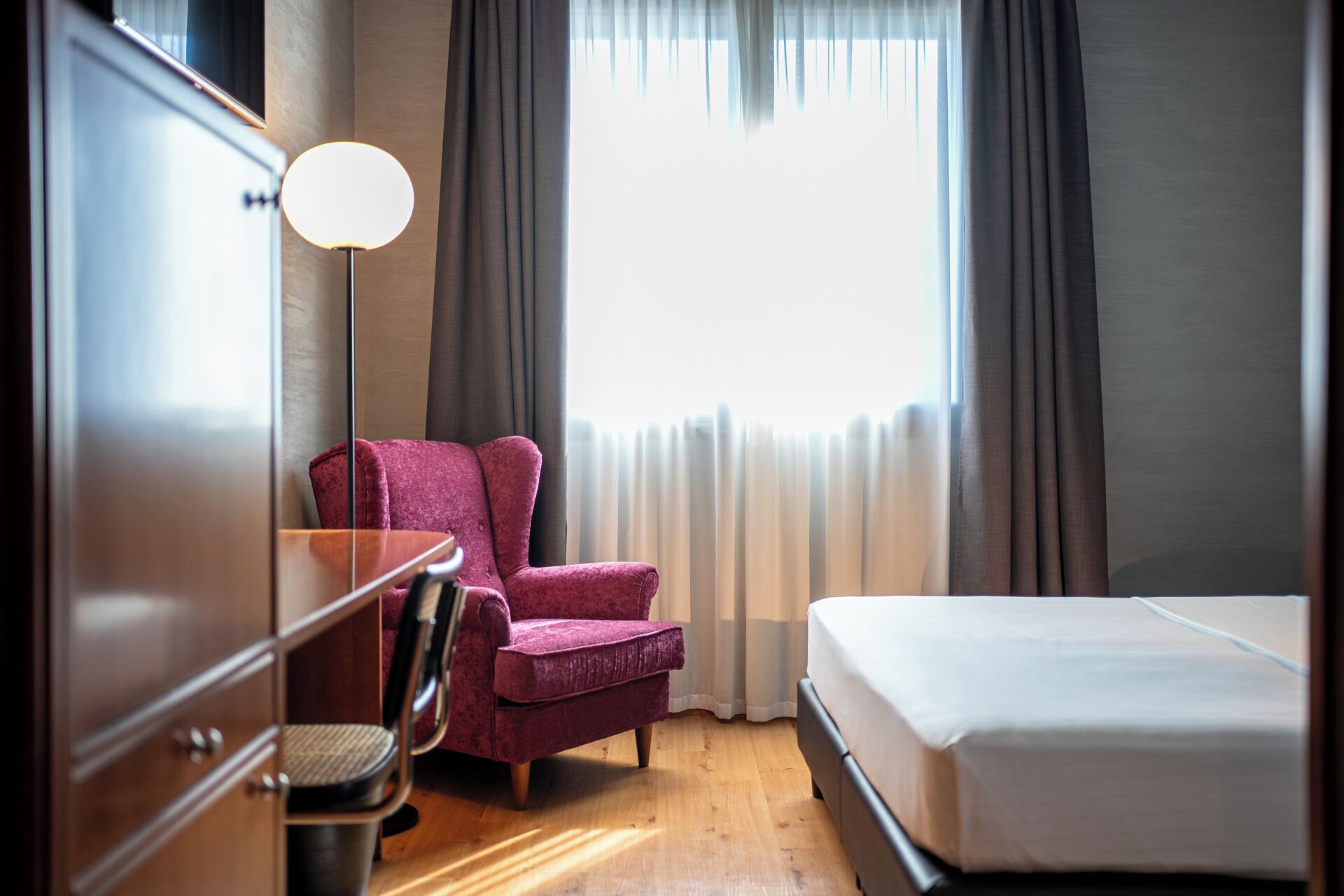 A hotel room with a bed, a desk with a chair, and a large window with curtains.