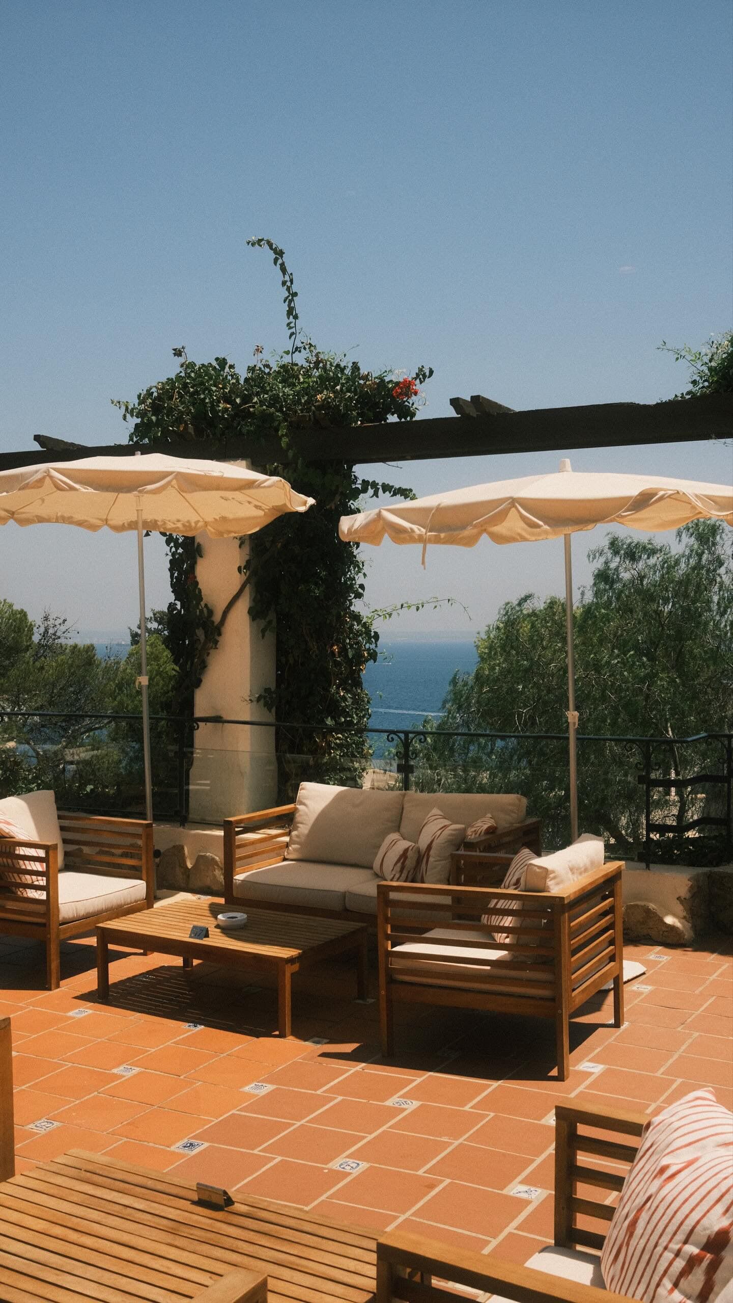 An outdoor seating area with wooden furniture, white umbrellas, and a view of the sea.