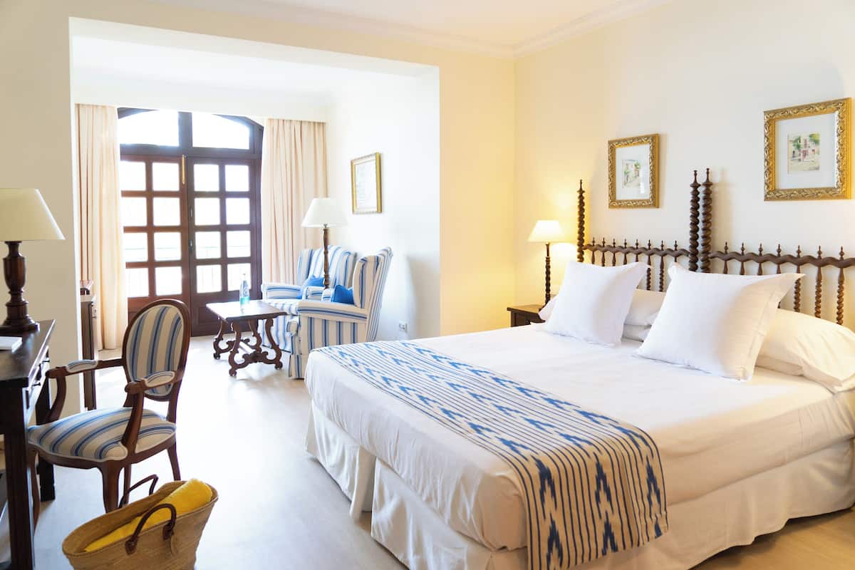 A hotel room with a large bed, a desk with a chair, a window with curtains, and framed pictures on the wall.
