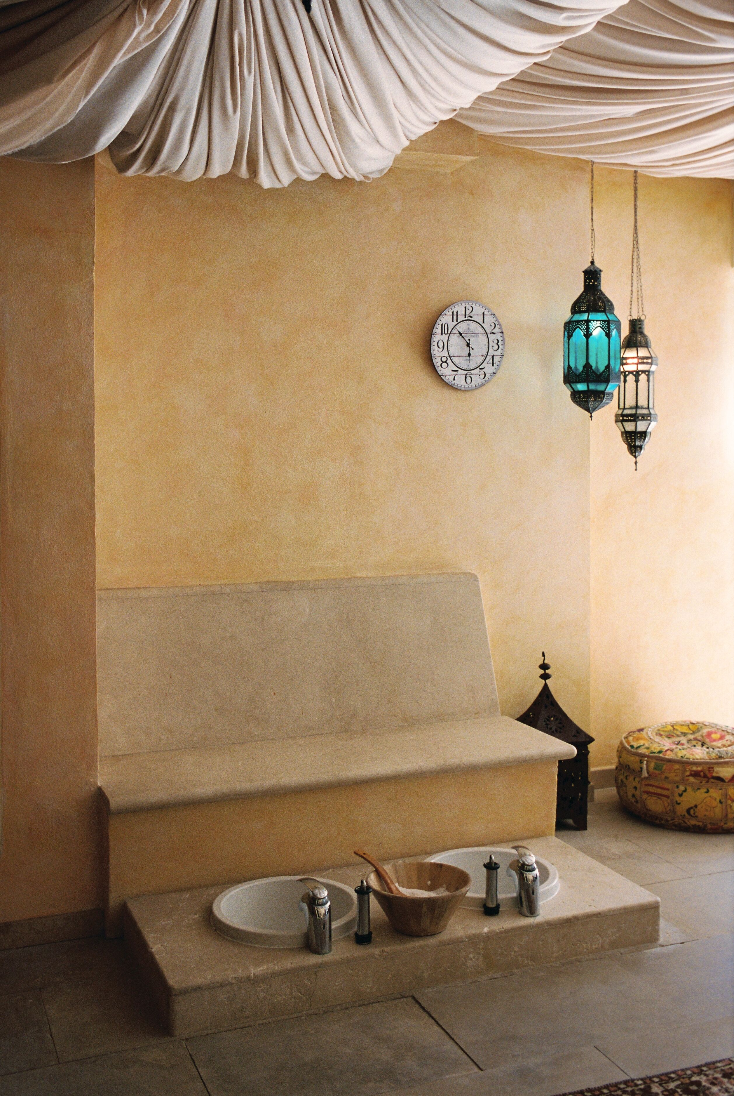 A beige sofa, a wall clock, two hanging lanterns, a small table with a bowl and a pitcher, and a decorative item on the floor.