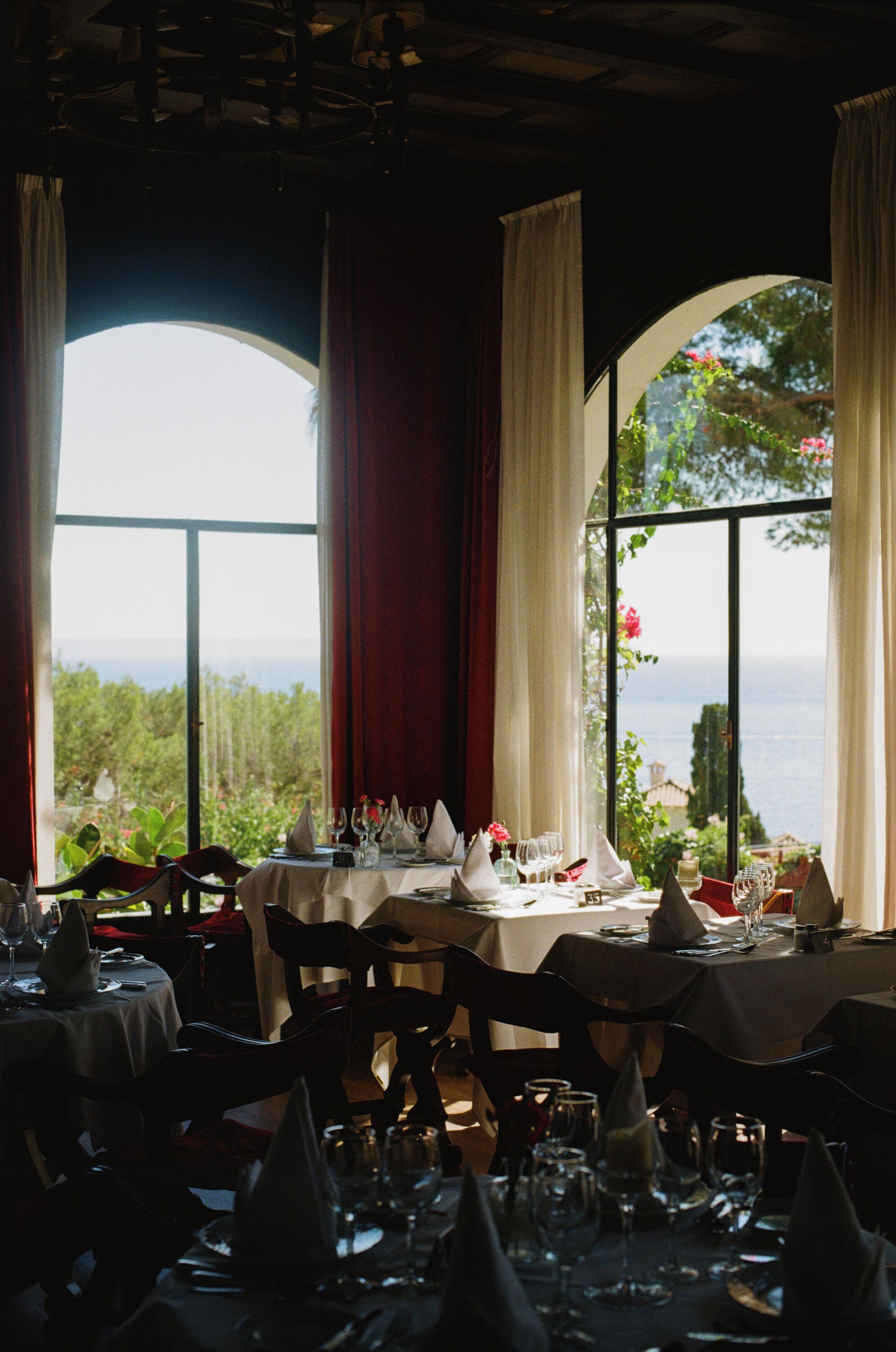 Un espace repas avec des tables dressées pour le repas, de grandes fenêtres avec des rideaux et une vue sur la verdure et la mer.