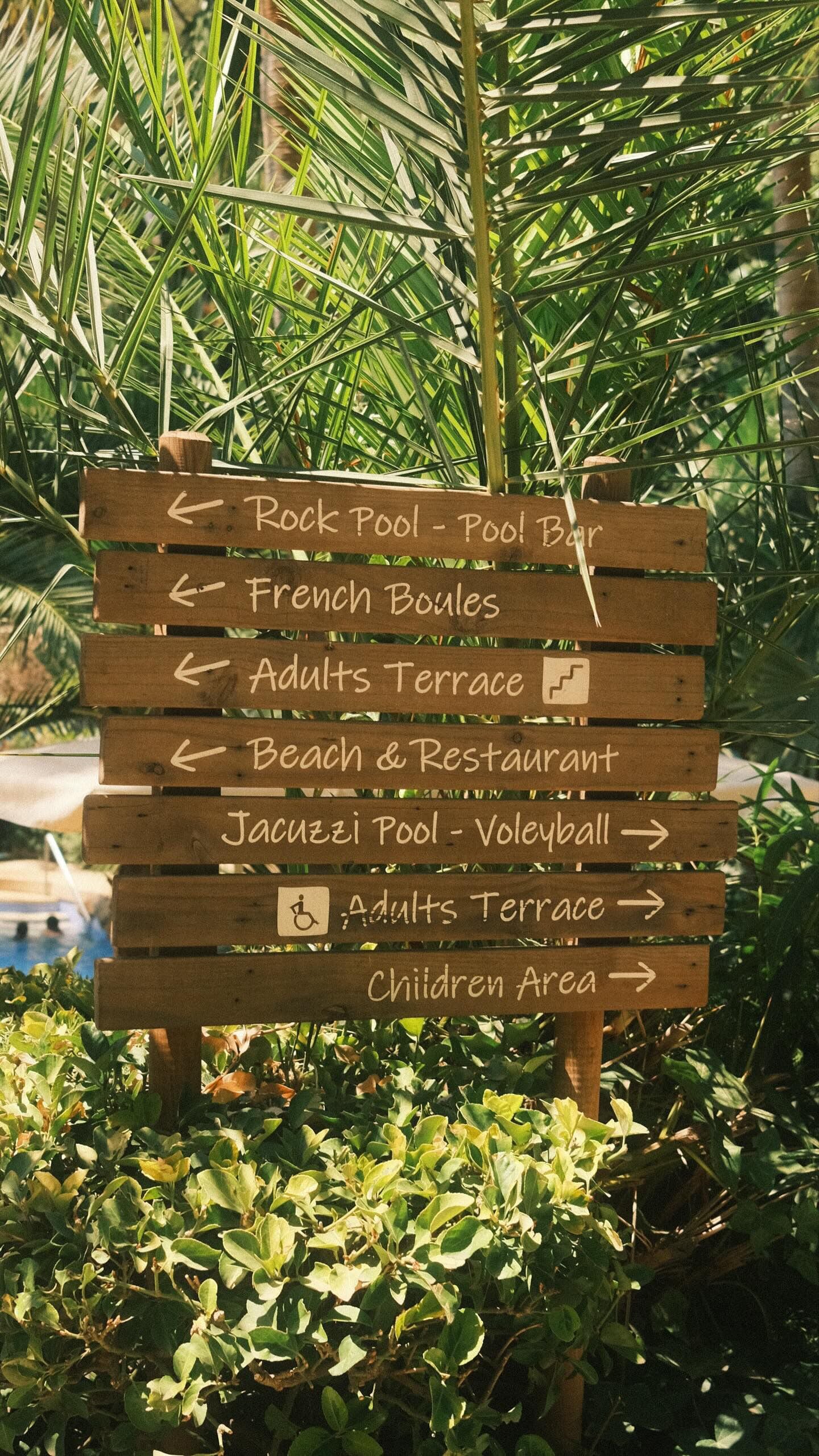 A wooden signpost with directional arrows pointing to various amenities such as Rock Pool, French Boules, Adults Terrace, Beach & Restaurant, Jacuzzi Pool, Volleyball, and Children Area.