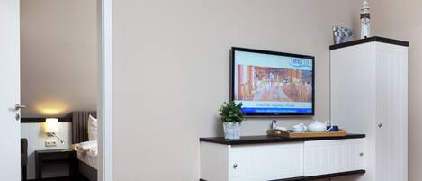 A hotel room with a television displaying a program, a bed, a nightstand with a lamp, a small plant, and a cabinet with a tray on top.