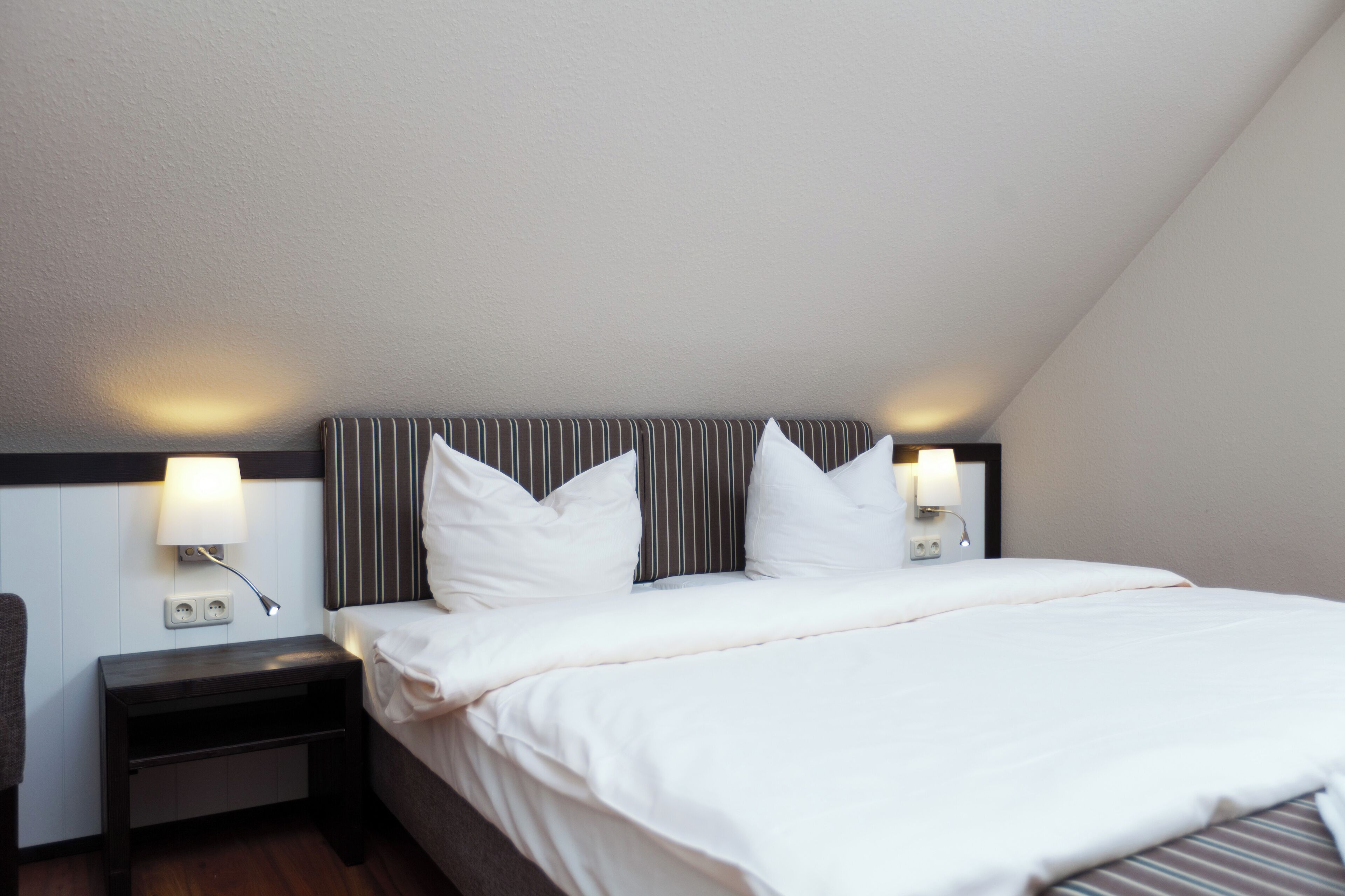 A hotel room with a bed, two bedside lamps, a nightstand, and a wall socket.