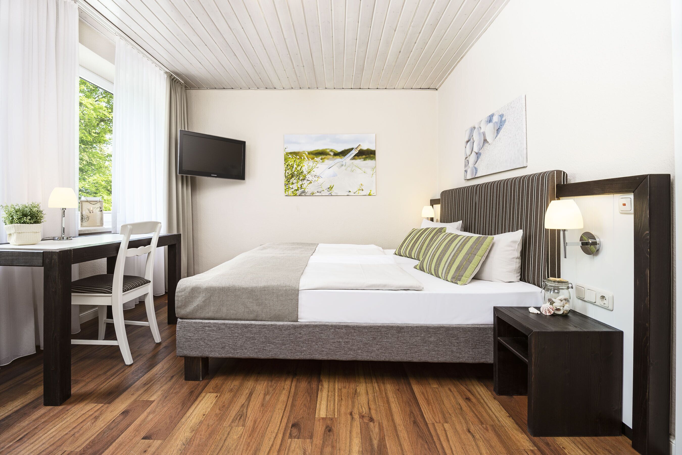 A bedroom with a bed, bedside tables, a desk, a chair, a TV, and artwork on the walls.