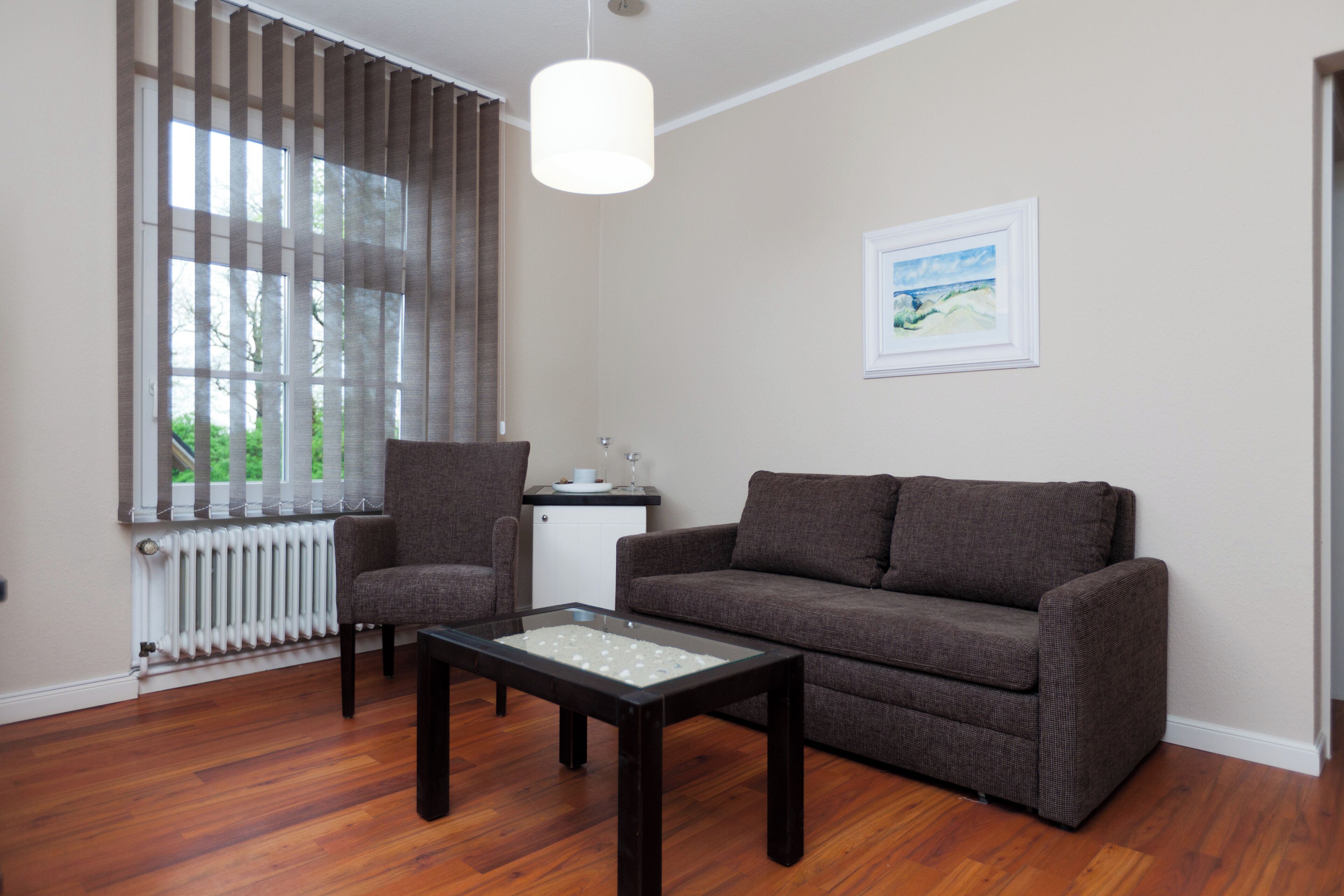 Ein Wohnzimmer mit Sofa, Sessel, Couchtisch, Heizkörper und einem fenster mit Vorhängen.