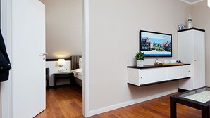 Quarto de hotel com televisão na parede, uma mesinha com tampo de vidro, uma cadeira e janela com vista para edifícios.