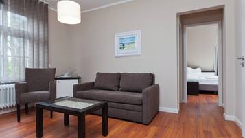 A room with a sofa, two armchairs, a coffee table, a painting on the wall, and a bed in the adjacent room.