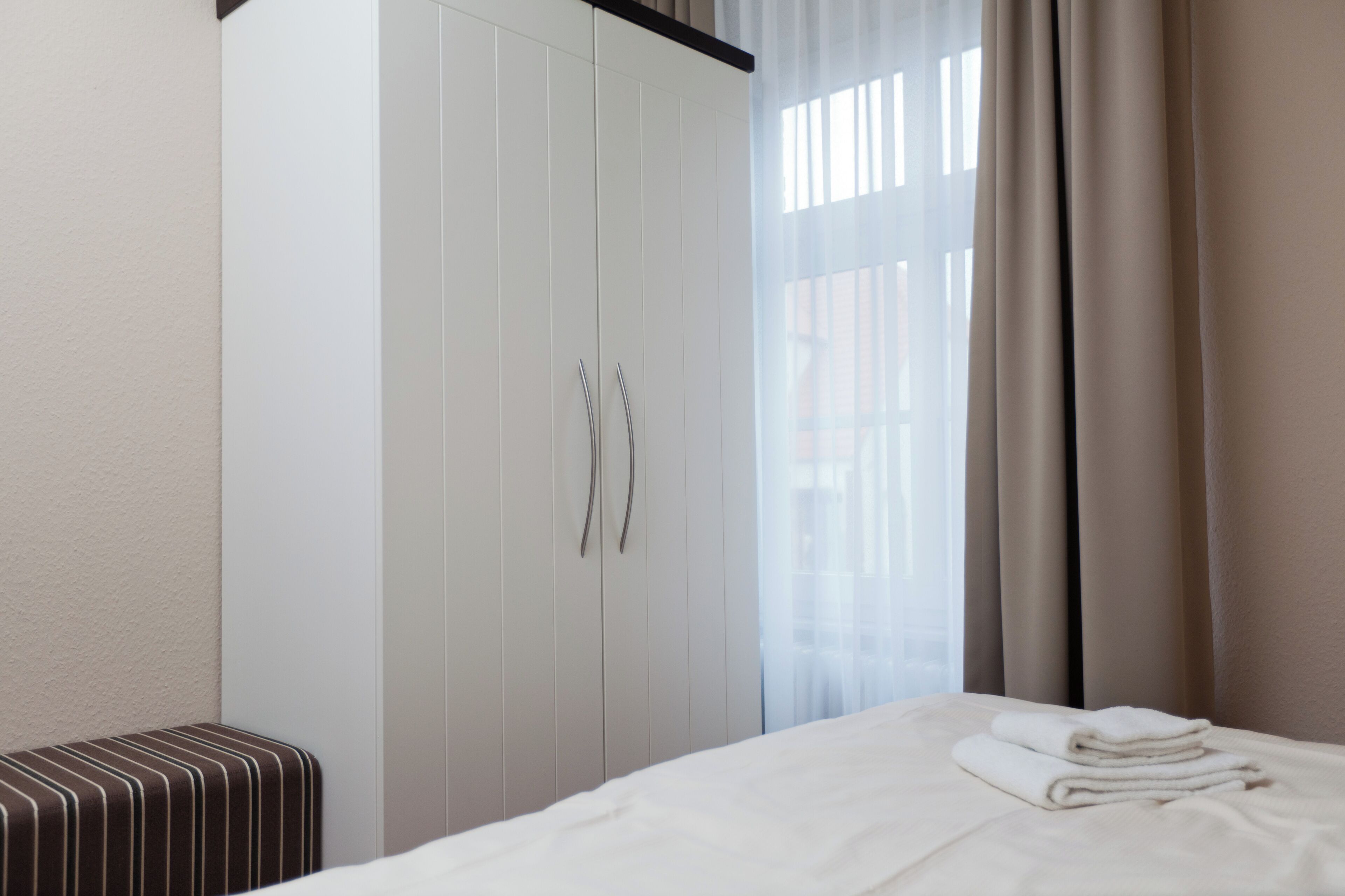 A hotel room with a bed, a wardrobe, a bench, and a window with curtains.