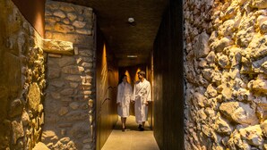 A stone-walled corridor with two individuals in white robes walking down. - Balneari Termes Victòria (Caldes de Montbui)