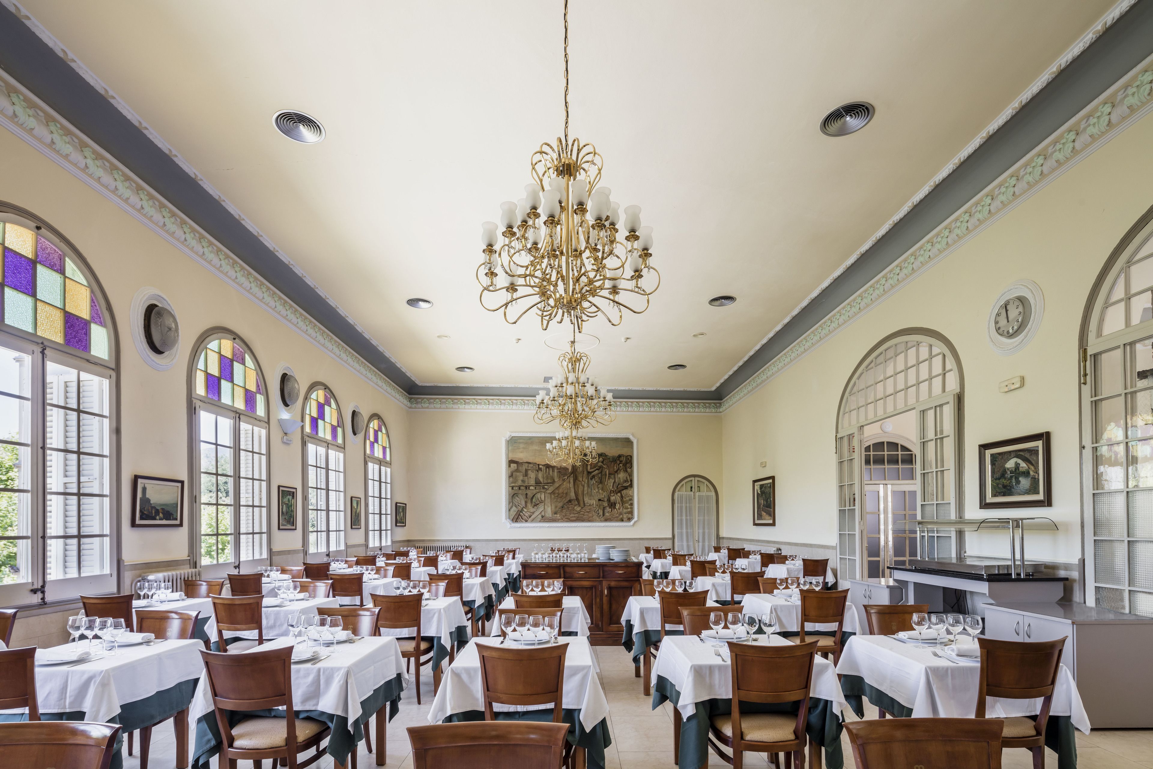 Une salle à manger spacieuse, dotée de nombreuses tables, d’élégants lustres et de grandes fenêtres.