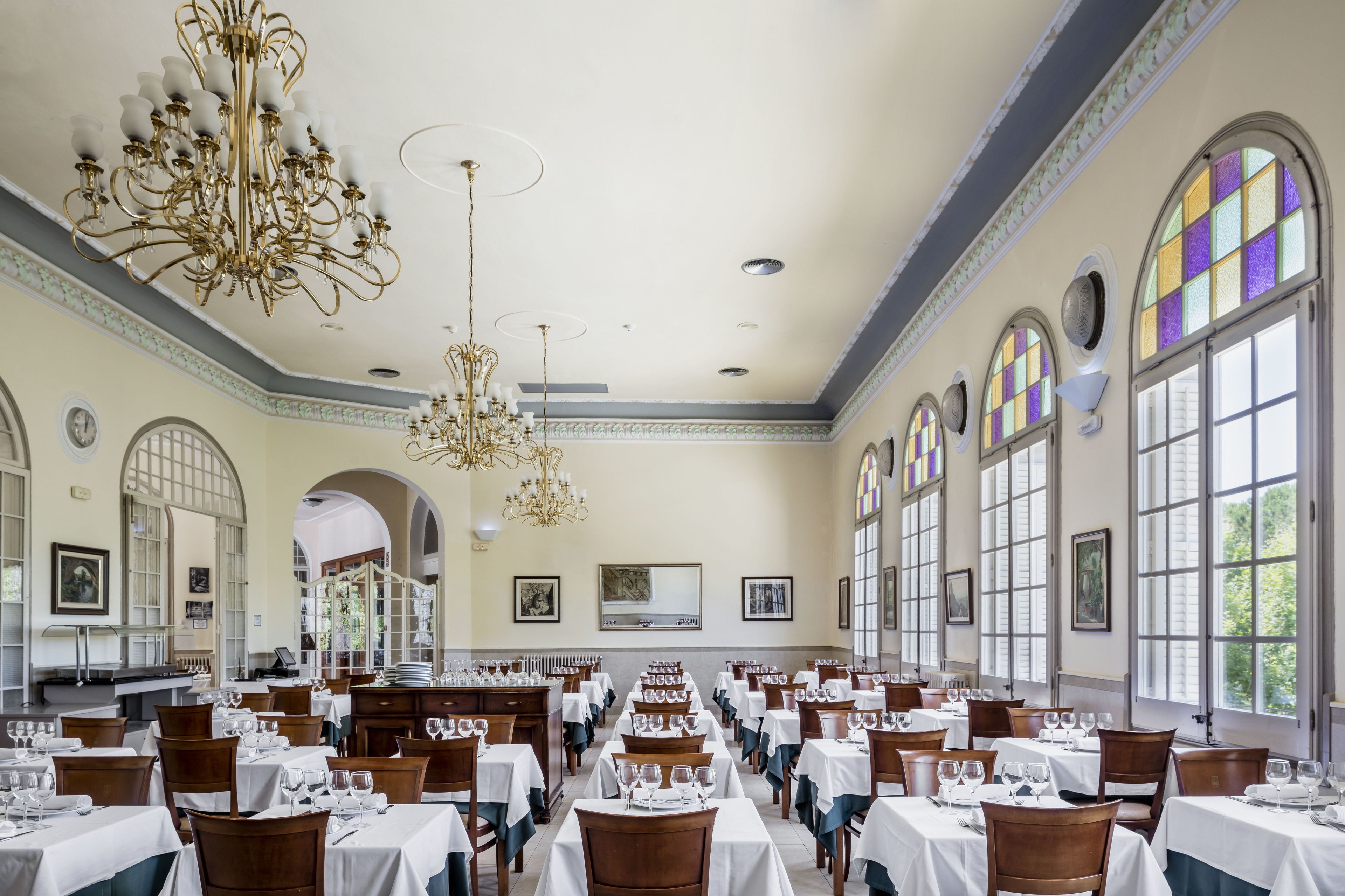 A spacious dining hall with multiple tables set for dining, large windows with stained glass, and ornate chandeliers.