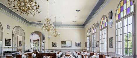 A spacious dining hall with multiple tables set for dining, large windows with stained glass, and ornate chandeliers.
