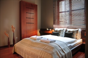 A neatly made bed with a beige blanket and two folded towels, a wooden wardrobe, and a window with blinds. - La Gioia Modern Designed Studios (Kraków)