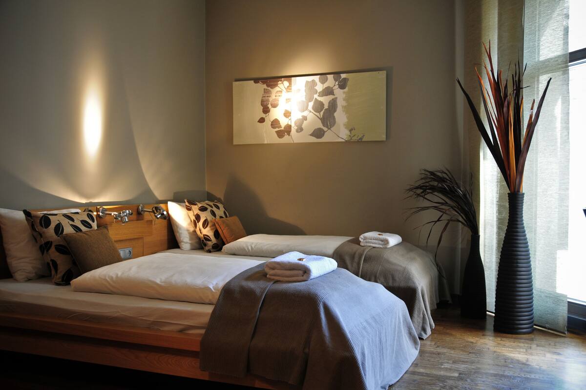 Un dormitorio con una cama grande, una mesita de noche, una obra de arte colgada en la pared y un jarrón alto con ramas decorativas.