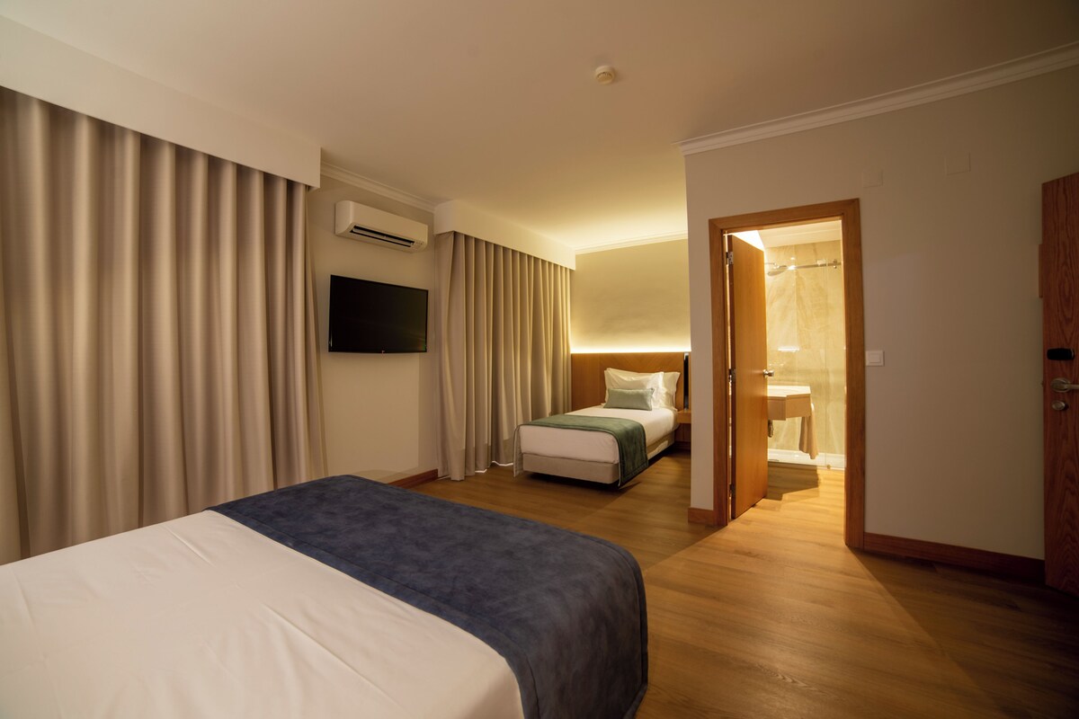 A hotel room with a bed, a TV, a bedside table, a window with curtains, and a bathroom visible through an open door.
