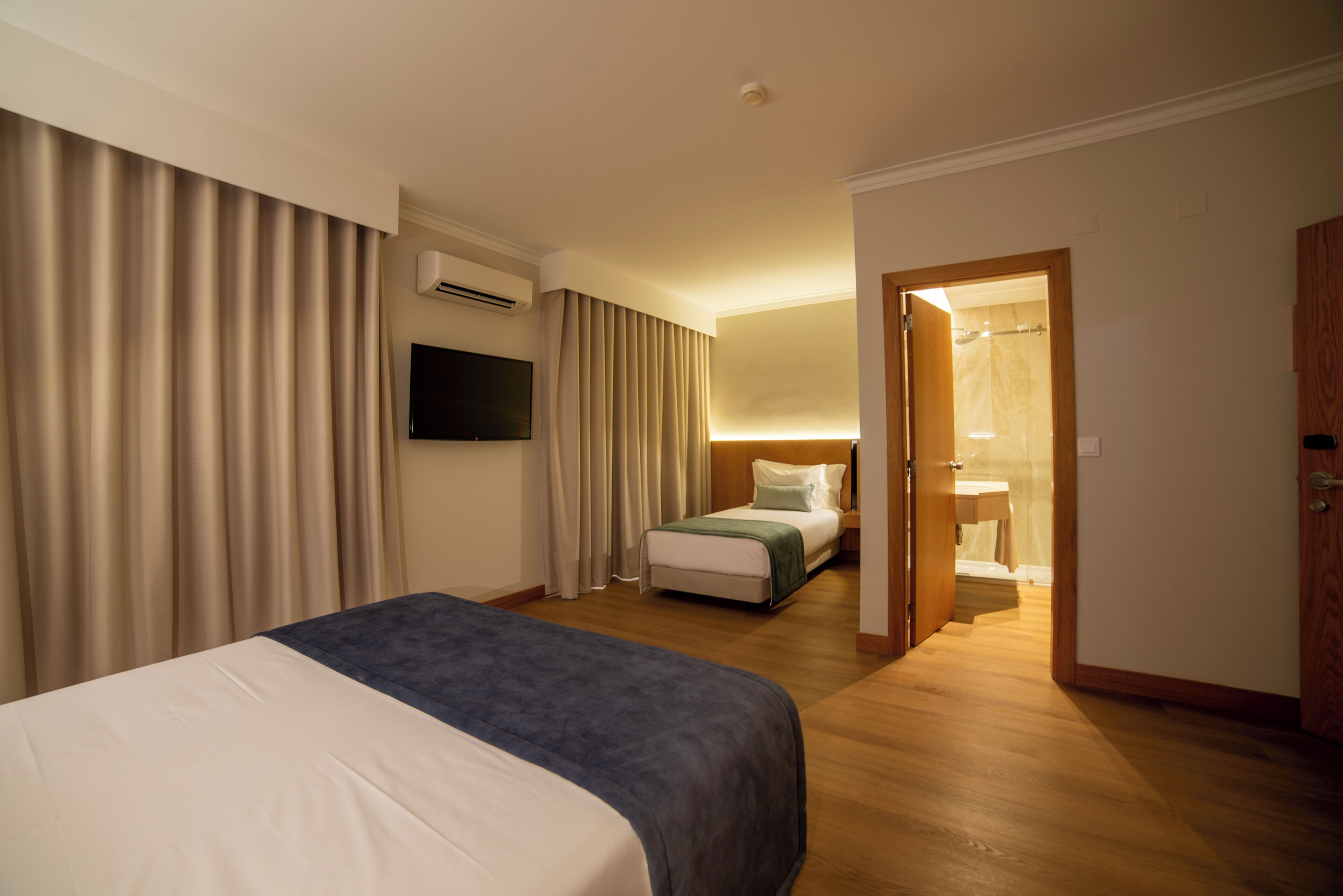 A hotel room with a bed, a TV, a bedside table, a window with curtains, and a bathroom visible through an open door.