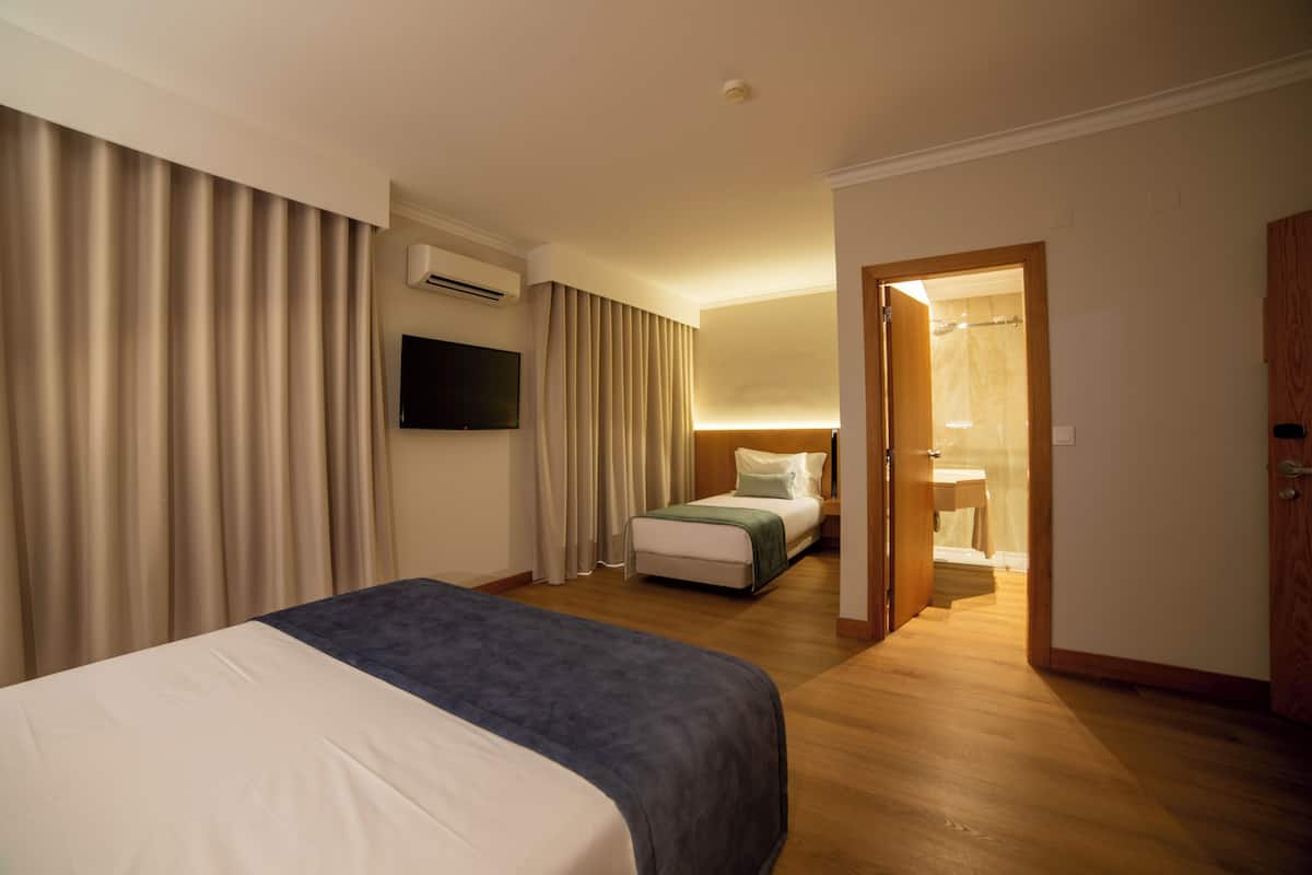 Habitación de hotel con cama, televisor, mesita de noche, ventana con cortinas y baño visible a través de una puerta abierta.