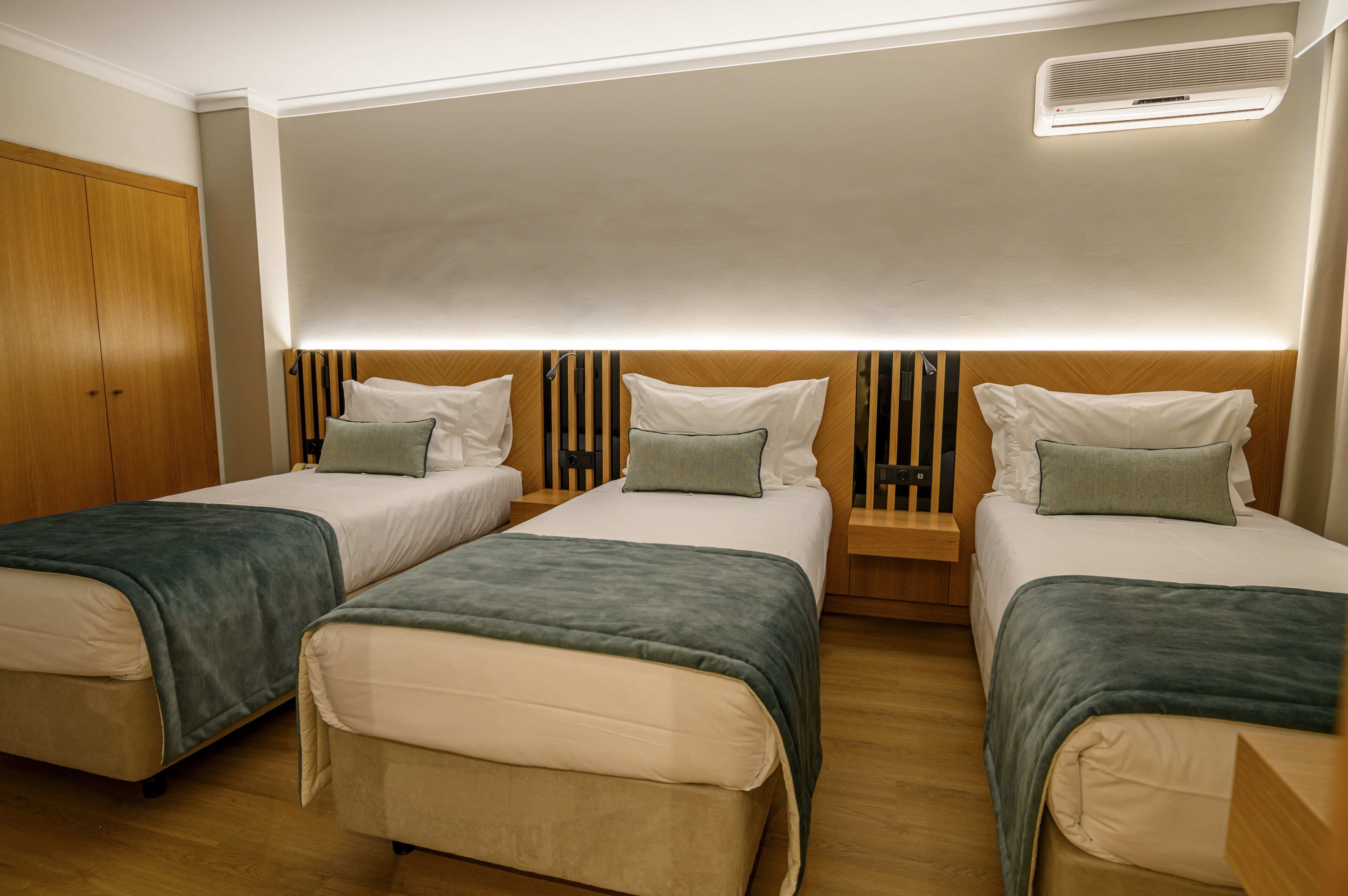 A hotel room with three single beds, wooden headboards, and a built-in air conditioning unit.