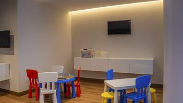 Ein Spielzimmer für Kinder mit einem weißen Tisch, bunten Stühlen, einem weißen Schrank und einem an der Wand montierten Fernseher.