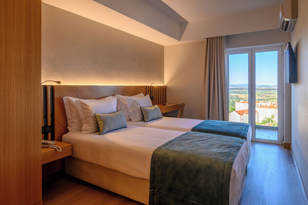 Habitación de hotel con una cama grande, una mesita de noche, una lámpara y vistas a través de un ventanal.