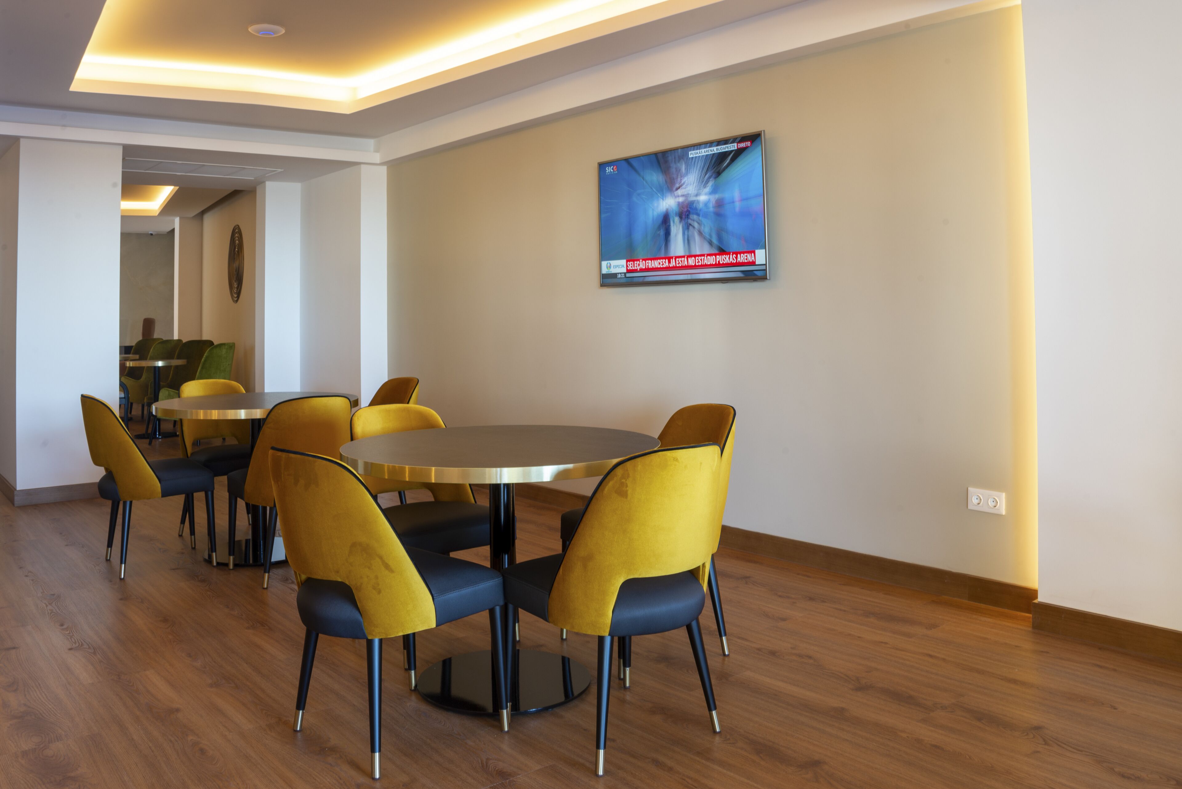 A modern dining area with round tables and yellow chairs, a flat-screen TV on the wall, and a wooden floor.