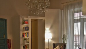 Un salon avec un lustre, une bibliothèque, un fauteuil, une table avec une lampe et une fenêtre avec des rideaux.