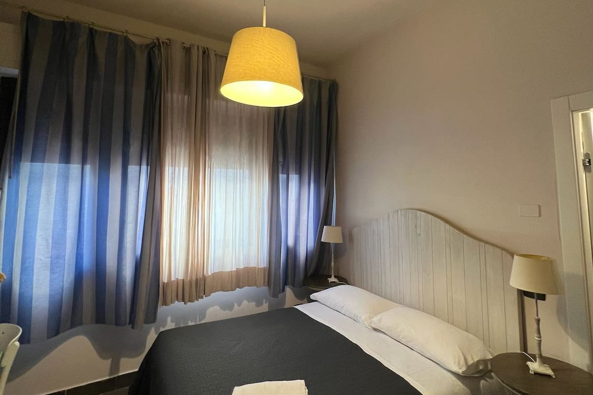 A hotel room with a bed, a bedside table, a lamp, and a window with curtains.