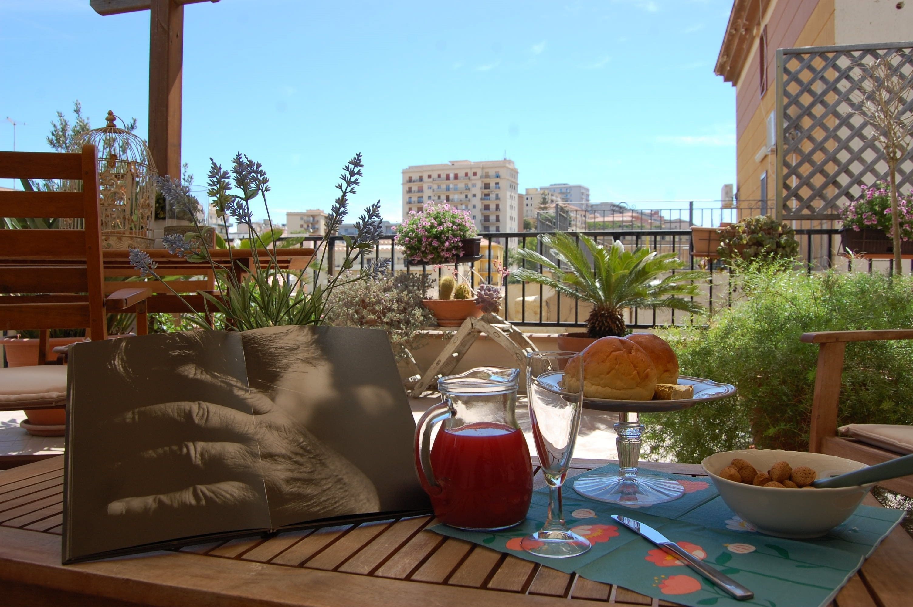 A table with a laptop, a pitcher of red drink, a glass, a plate with bread, and a bowl of snacks on a patio with plants and a city view.