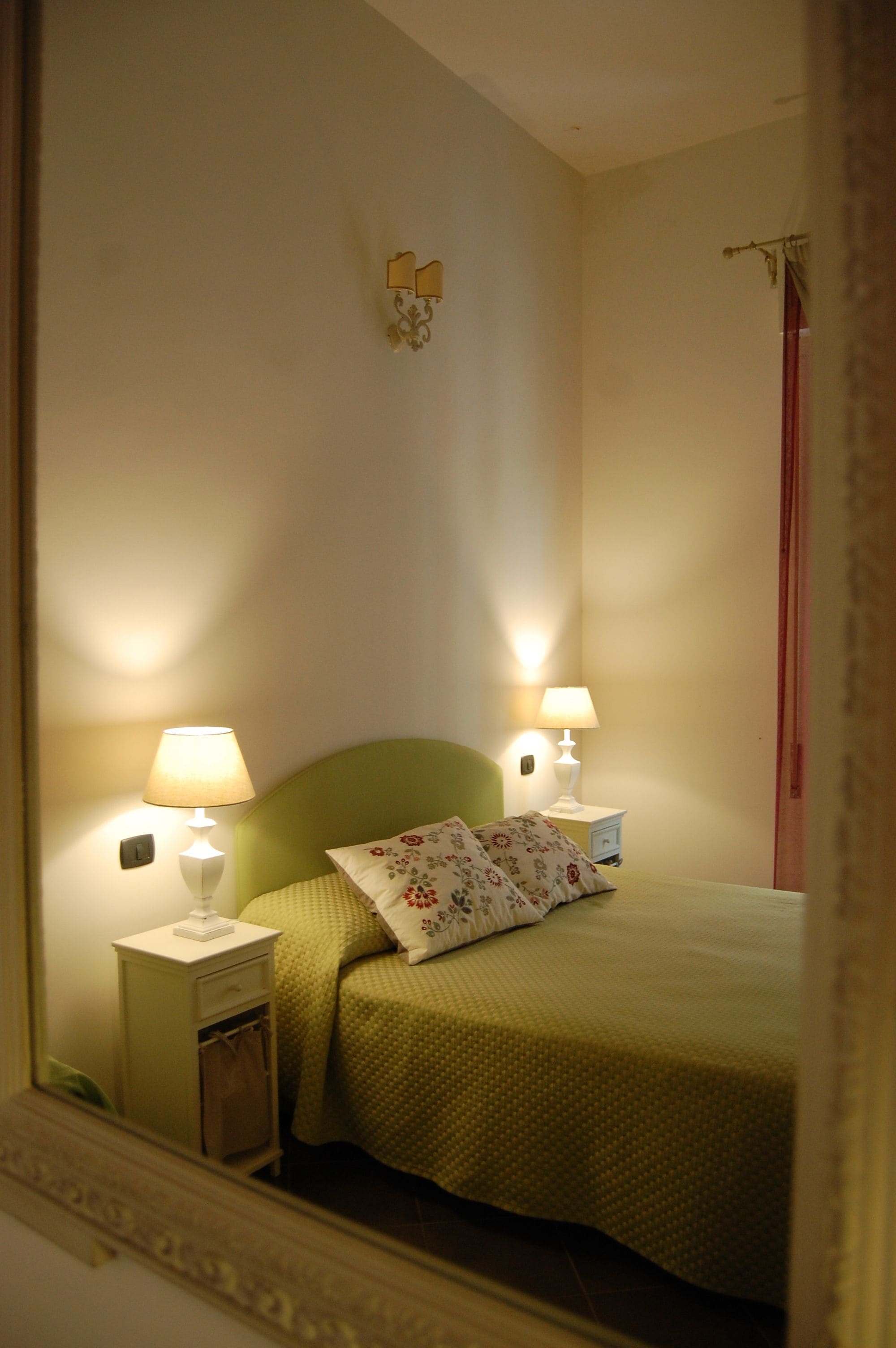 A bedroom with a green bed, two bedside lamps, a nightstand, and a mirror reflecting the room.