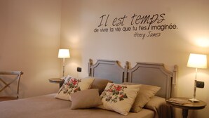 Un lit bien fait, agrémenté d’oreillers à motifs floraux, une table de chevet avec une lampe et une citation d’Henry James affichée sur le mur.
